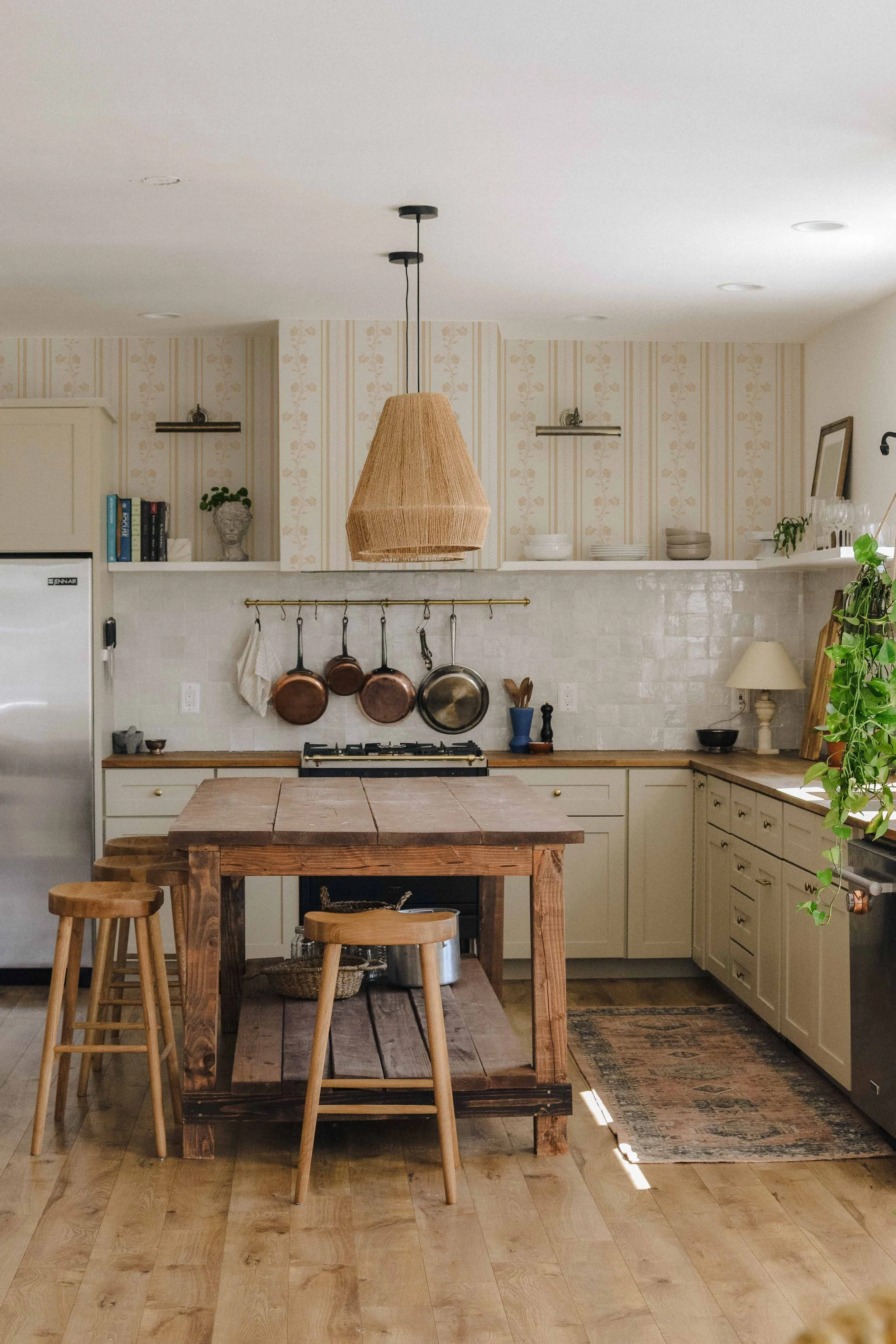A cozy kitchen with a rustic wooden dining table, two wooden stools, a hanging wicker lamp, white cabinets, a countertop with various kitchen items, a rug, and greenery.