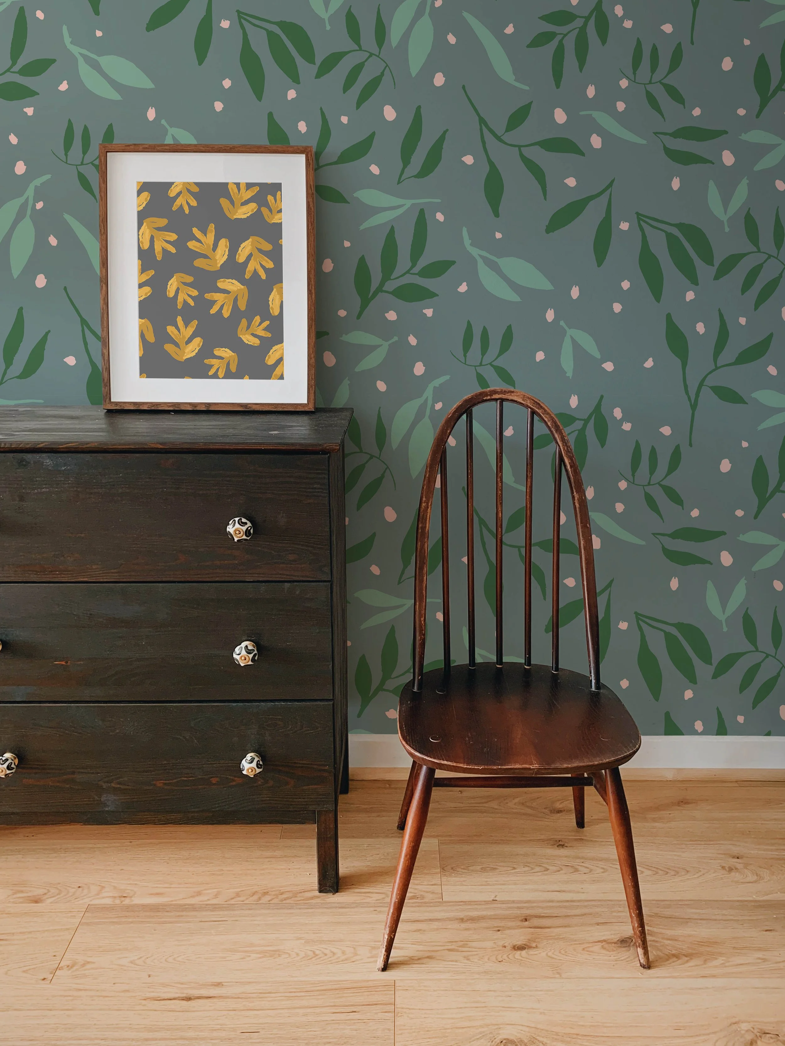 A dark wooden dresser with four drawers, each with decorative ceramic knobs, sitting on a light wood floor against a green, leafy patterned wallpaper. To the right of the dresser is a wooden chair with a curved backrest and four legs, also on the light wood floor.