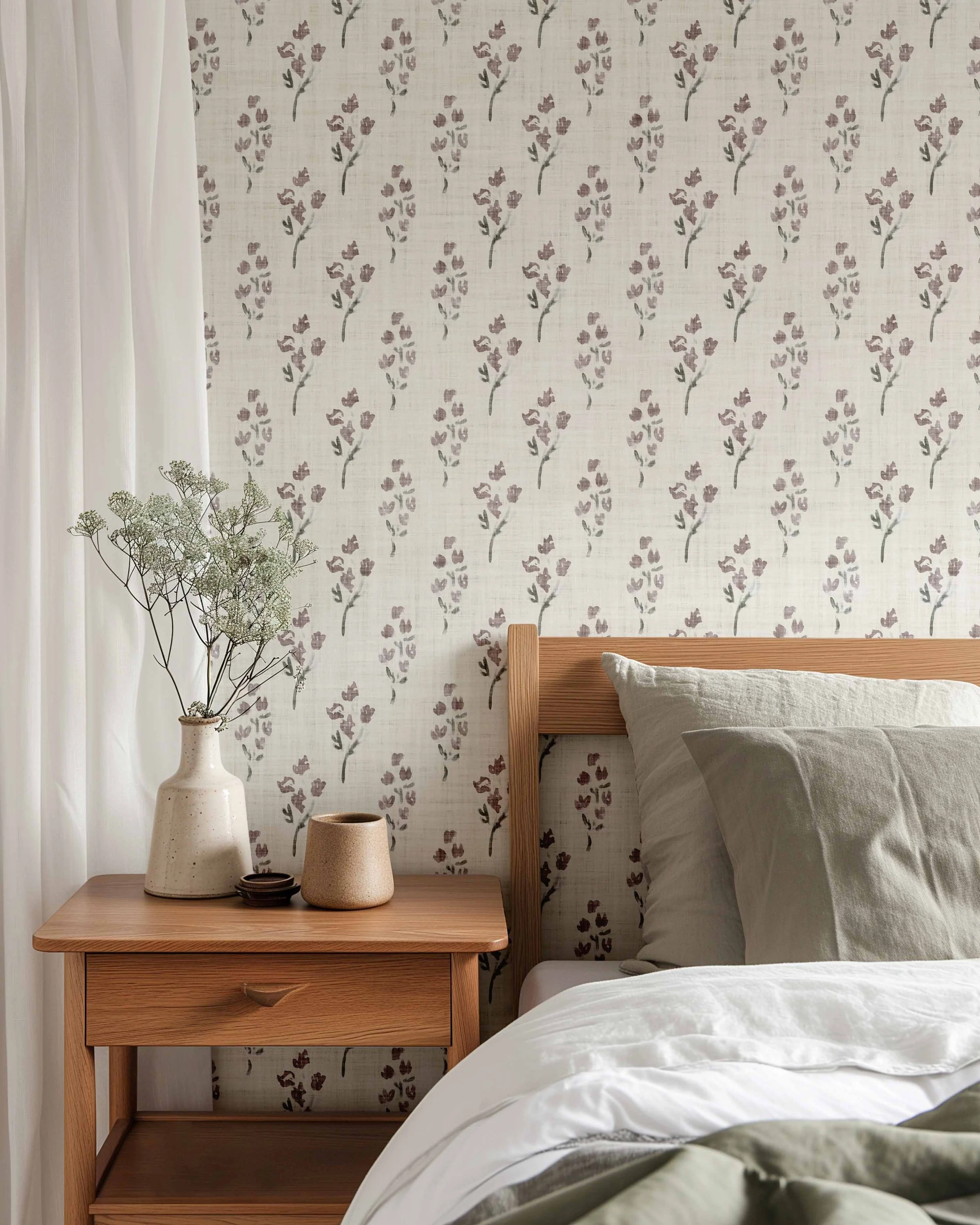 A cozy bedroom corner featuring a wooden nightstand with decorative pottery, a bed with white and beige linens, a floral wallpapered wall, and a plant near the window with white curtains.