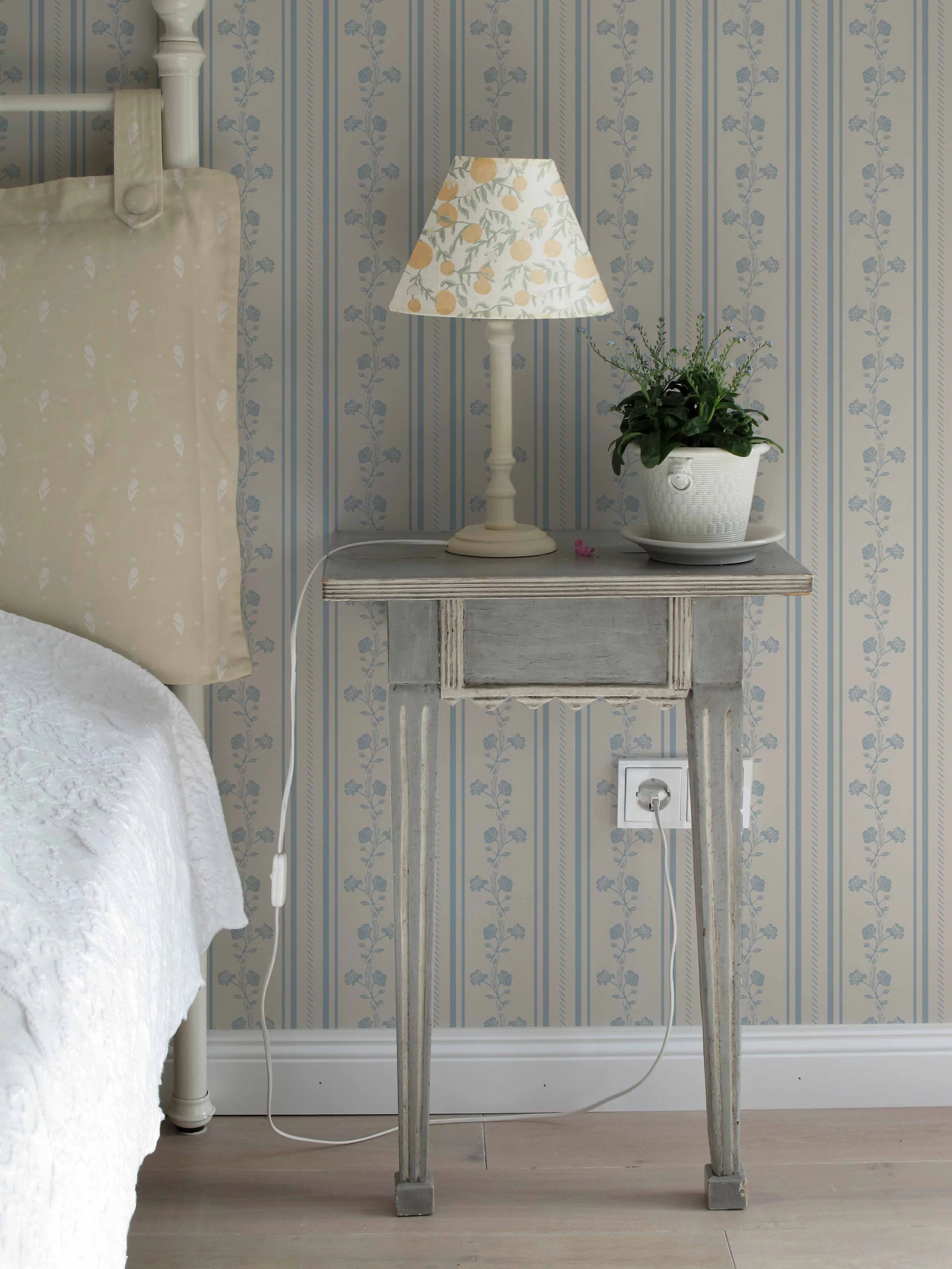 A beige bed headboard on the left side of the image with part of a white quilt visible. A wooden bedside table with a distressed finish, a white lamp with a patterned shade featuring yellow and green fruit and leaf design, a potted green plant in white basket-style pot, located on a light-colored wooden floor. An electrical outlet and a power plug are visible at the bottom of the image. The wall has blue and beige floral-patterned wallpaper.