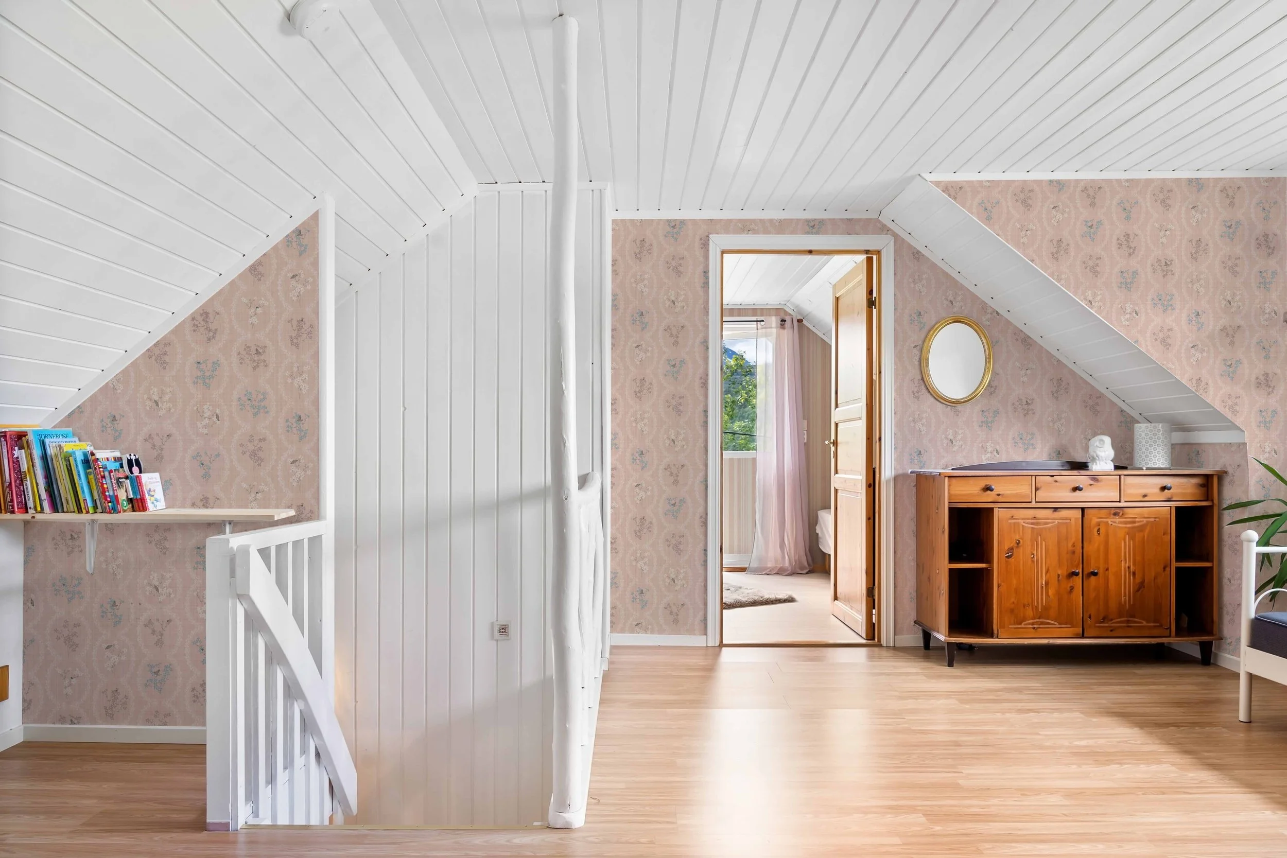 Interior of an attic room with wooden flooring, pink floral wallpaper, white-paneled ceiling, a bookshelf with children's books on a ledge, a wooden sideboard, a round mirror, a statue, a white speaker, and a bedroom visible through an open door with pink curtains and a window.