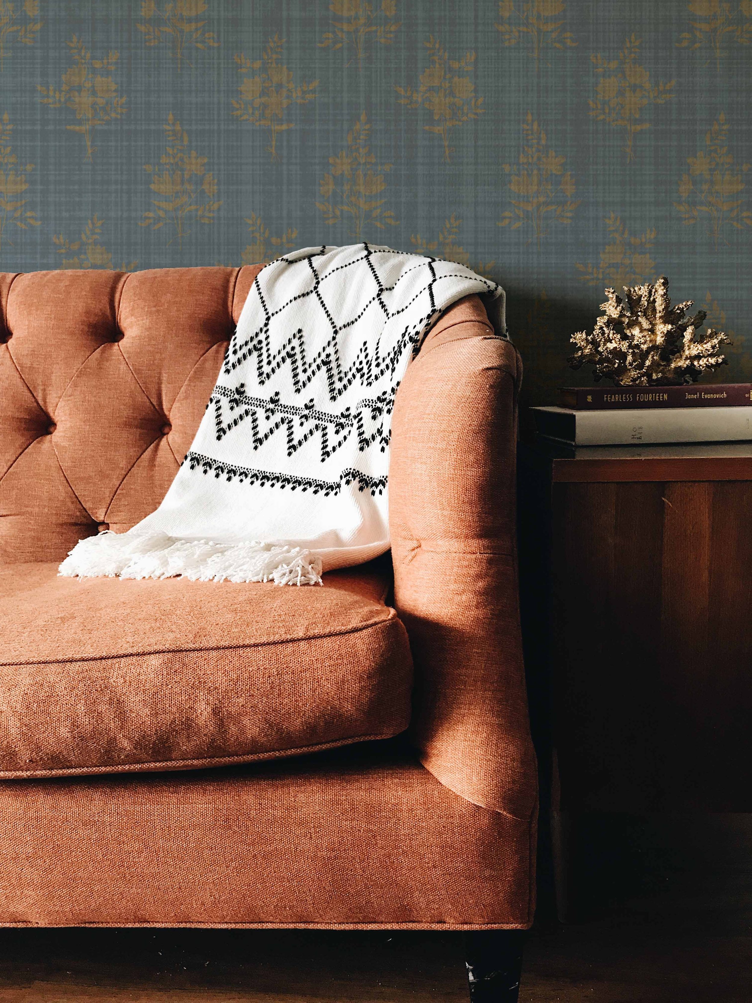 A cozy living room corner featuring a rust-colored tufted sofa with a white and black patterned throw draped over the backrest. To the right, a dark wooden side table holds a small decorative piece resembling dried coral and a few stacked books.