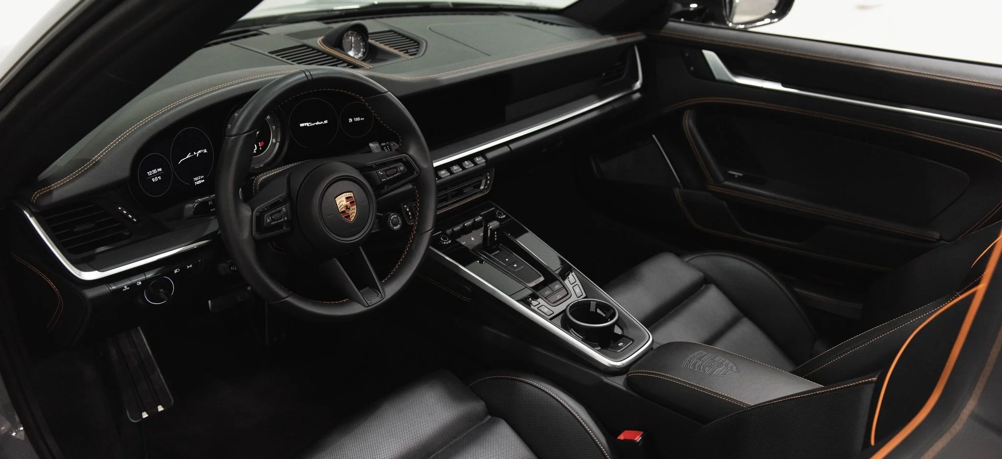 Interior of a black Porsche sports car, showing a steering wheel with the Porsche emblem, digital dashboard displays, center console with gear shifter and controls, black leather seats with orange stitching, and a modern dashboard with a mounted analog clock.