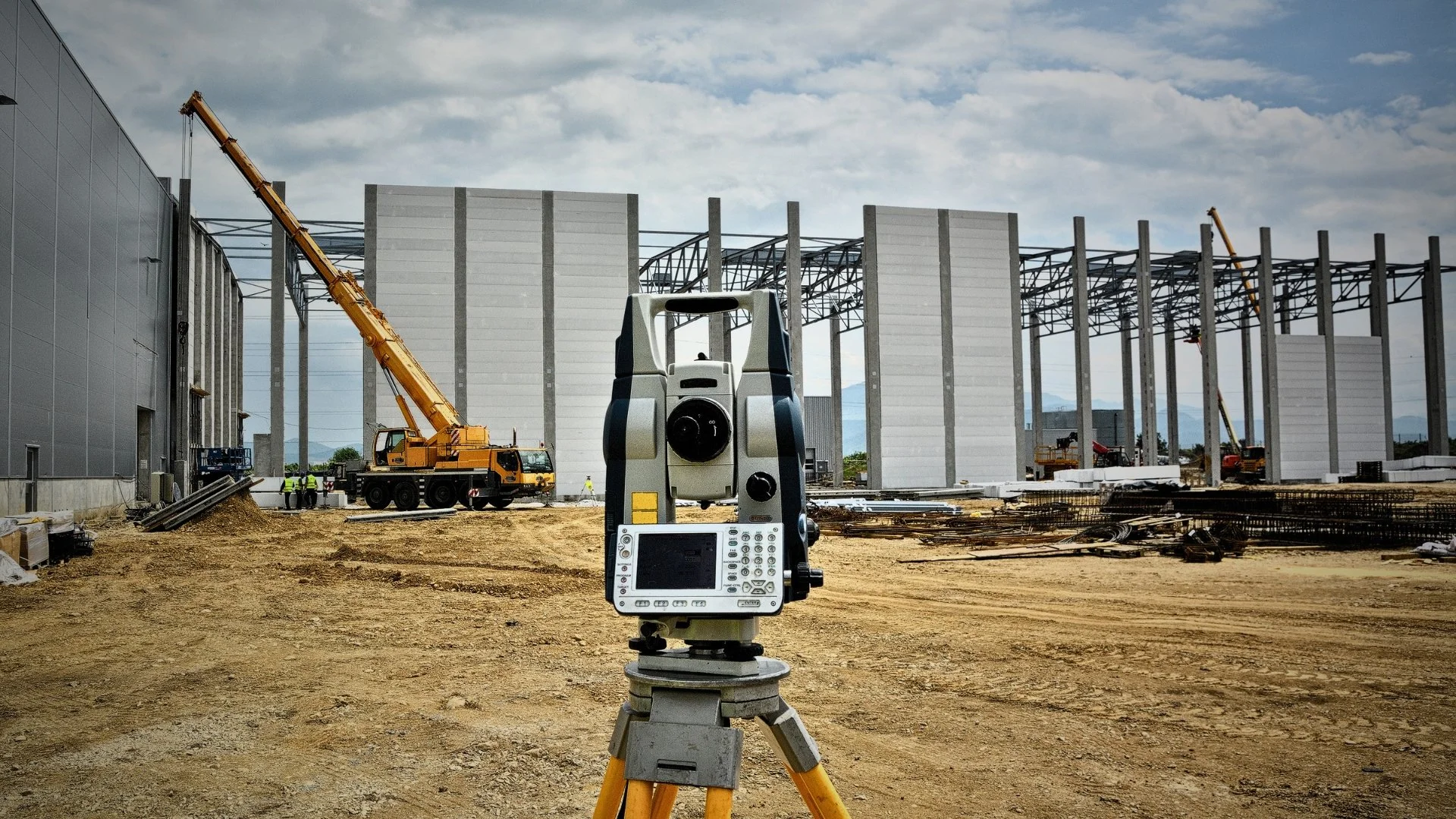 Construction site with a theodolite in the foreground, cranes lifting large panels, and partially built steel framework for a new building against a cloudy sky.