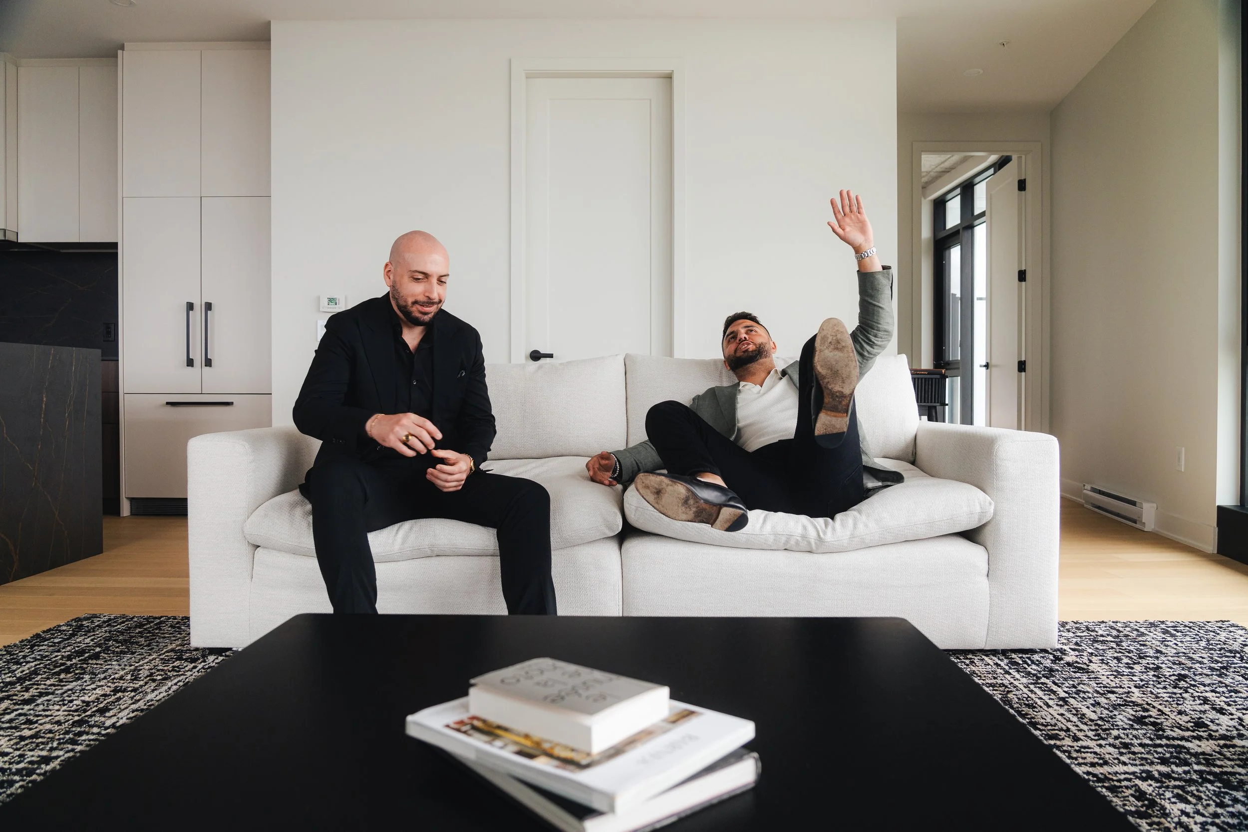 Two men sitting in a living room on a white couch, one flipping through a magazine on a black coffee table, the other lying back with one arm raised.