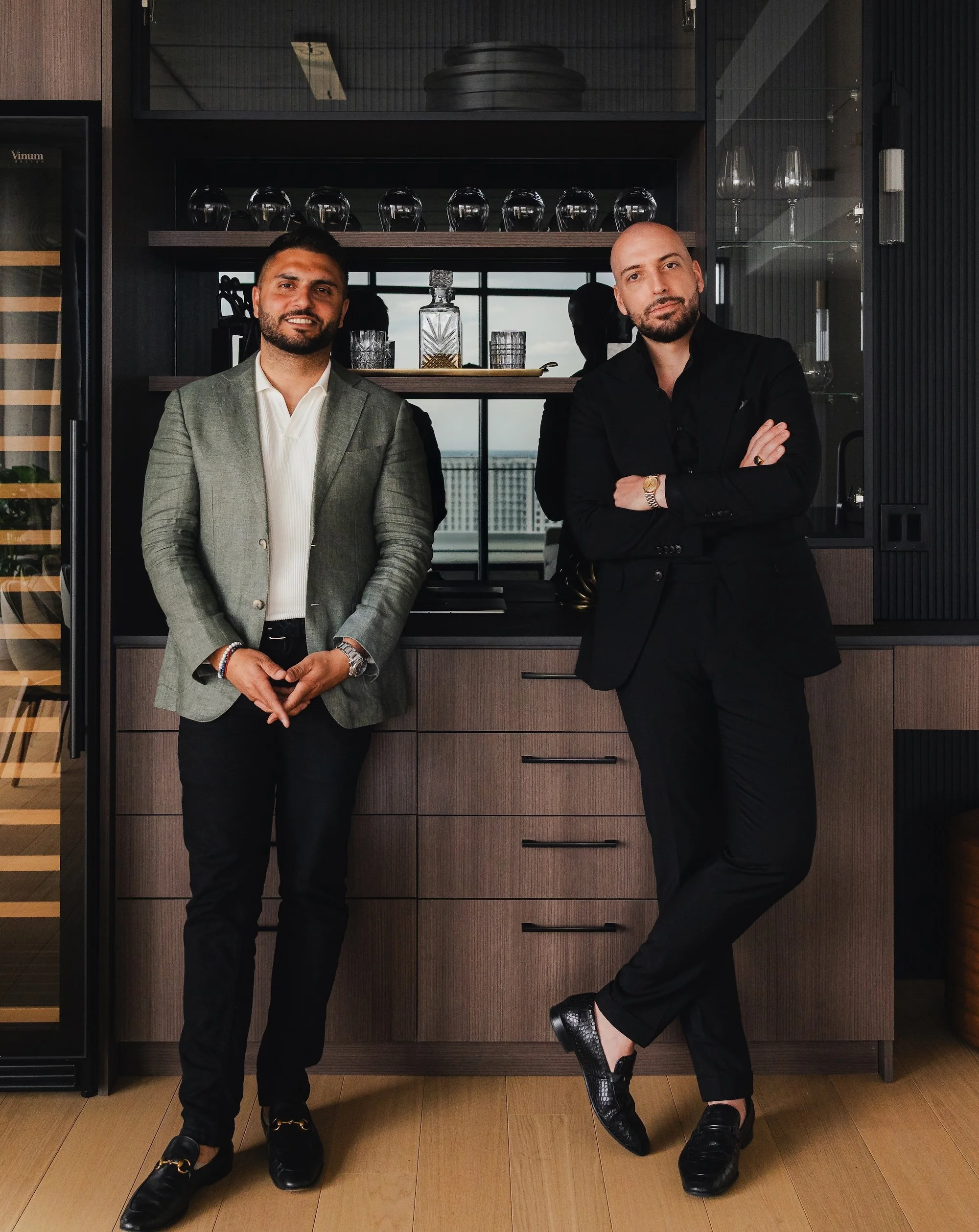Two men in formal attire posing indoors with a kitchen or bar backdrop featuring glassware and decorative items.
