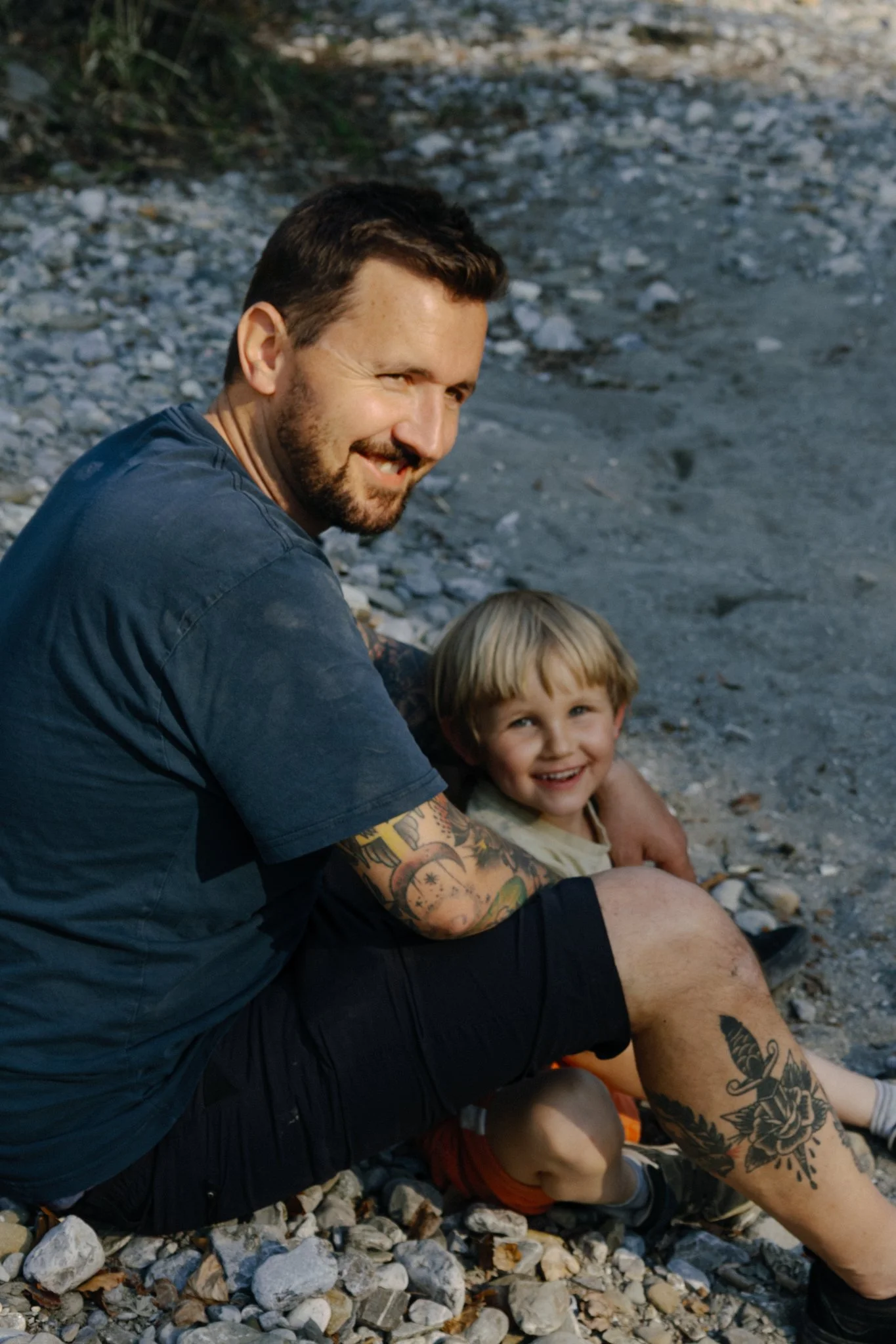 family photoshoot - A smiling man with tattoos sitting on a rocky beach with a young boy who also has tattoos. They are sitting closely together, and the man is looking at the camera while the boy is looking slightly away.