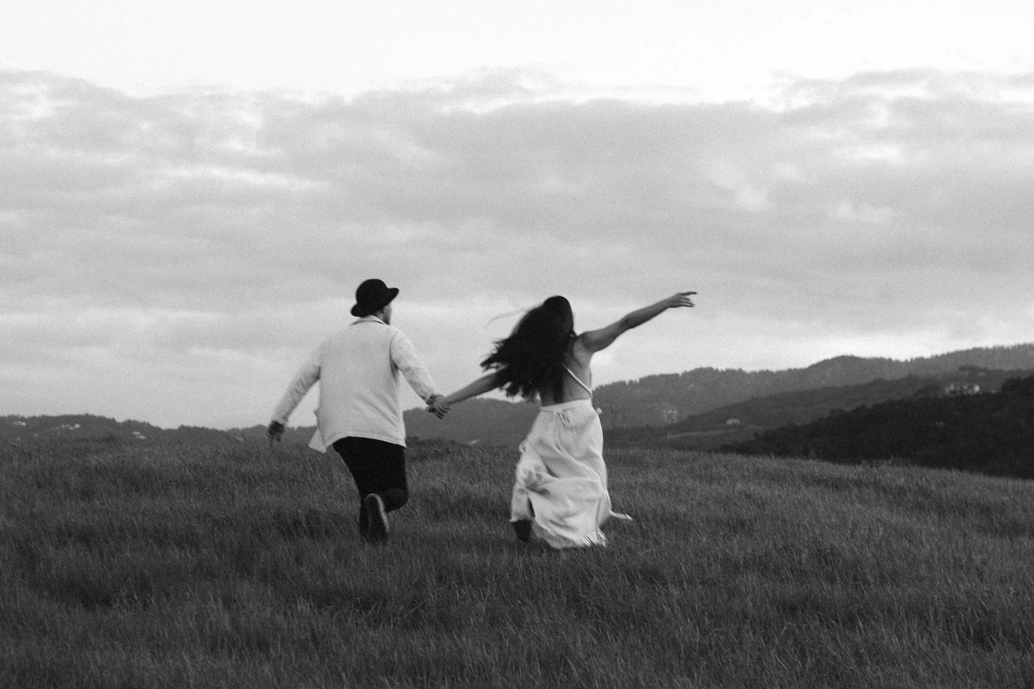 couples photoshoot - A black and white photograph of a man and woman holding hands and running through an open grassy field, with hills and a cloudy sky in the background.