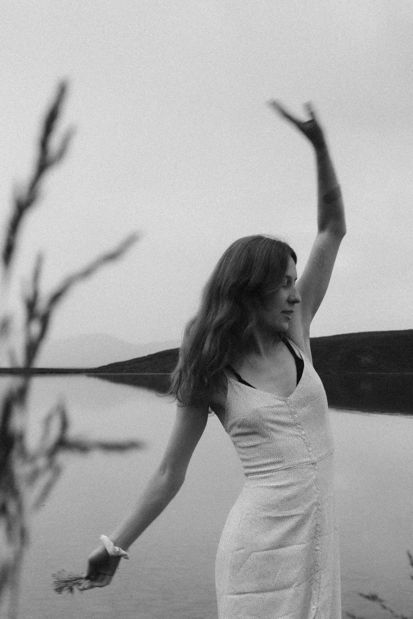 portrait - A woman with long hair standing outdoors by a body of water, wearing a sleeveless dress, with her arm raised, and a blurred plant branch in the foreground.