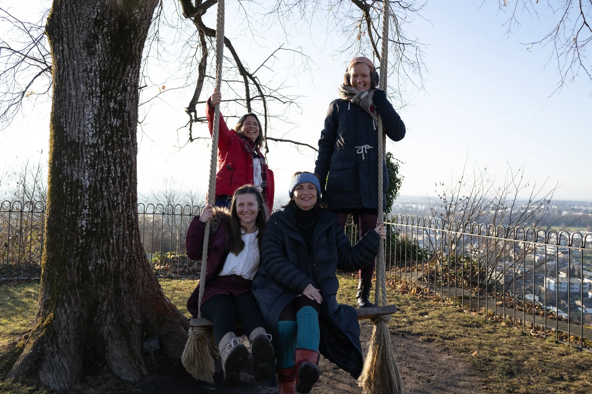 friends photoshoot - four women enjoying time on a wooden swing hanging from a large tree outdoors in a park, with city buildings in the background on a clear day.