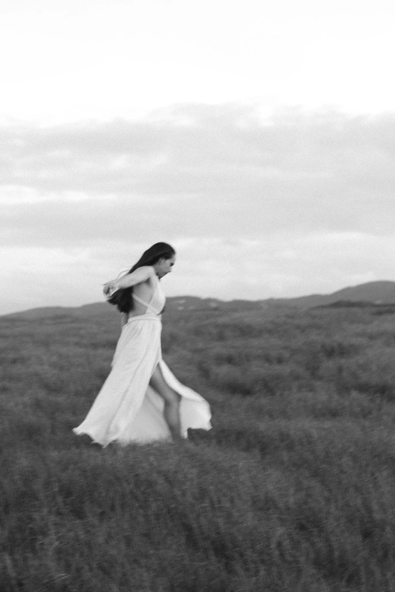 couples photoshoot - Black and white photo of a woman in a long dress walking on a grassy field with hills and cloudy sky in the background.