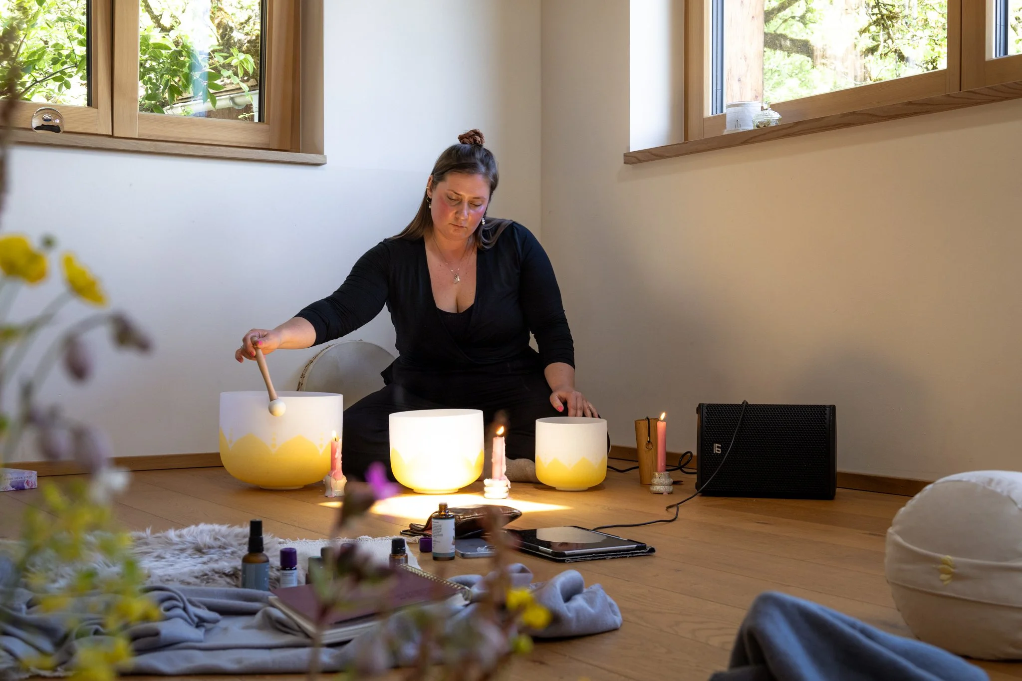 retreat photography - A woman is sitting on the floor in front of a row of singing bowls, candles, and essential oils, creating a calming atmosphere in a brightly lit room with windows showing a green outdoor scene.