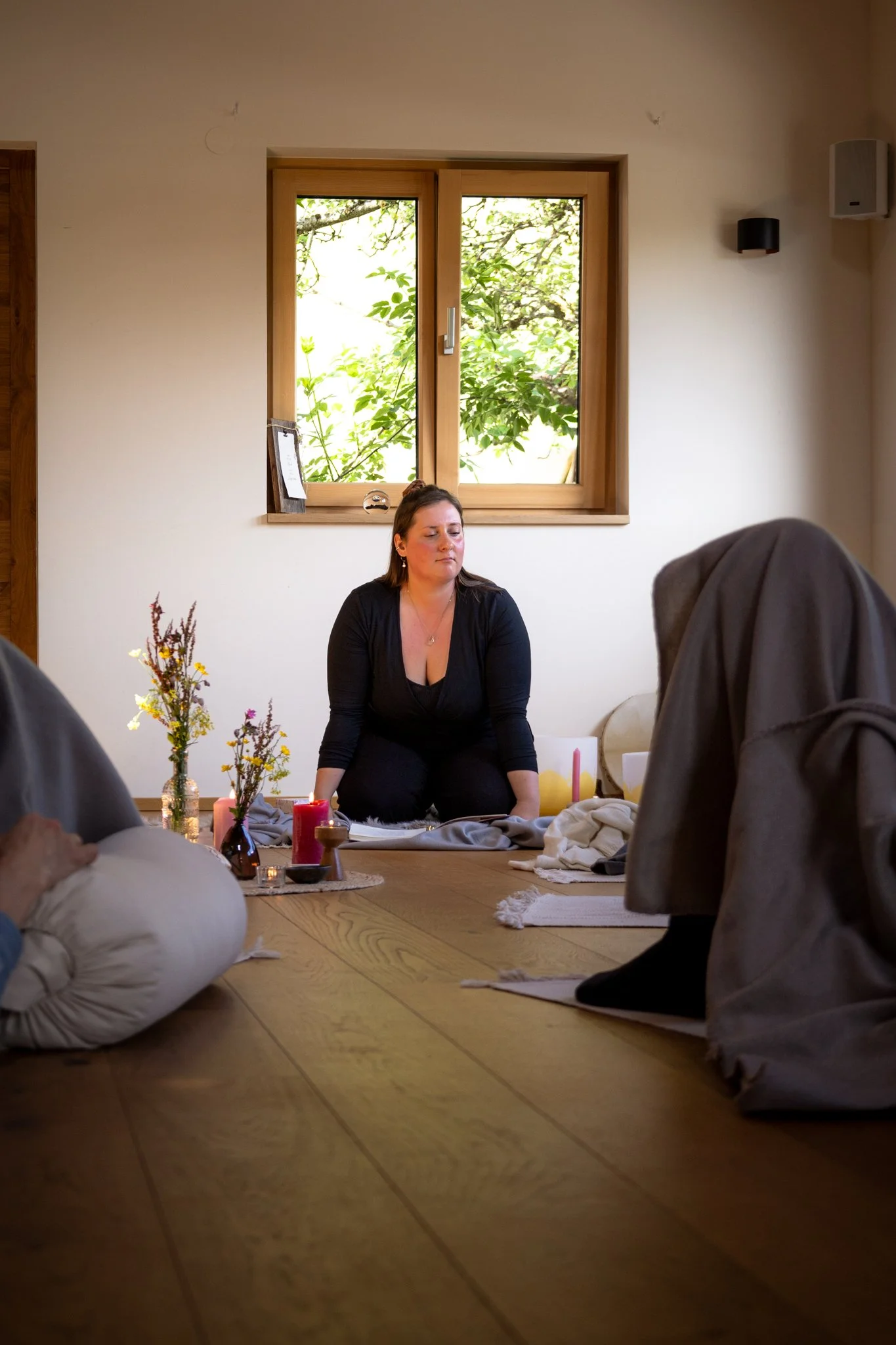 retreat photography - A woman sitting on the floor with her eyes closed during a meditation or prayer session, surrounded by candles, flowers, and other participants, in a cozy room with wooden window frames and natural light.