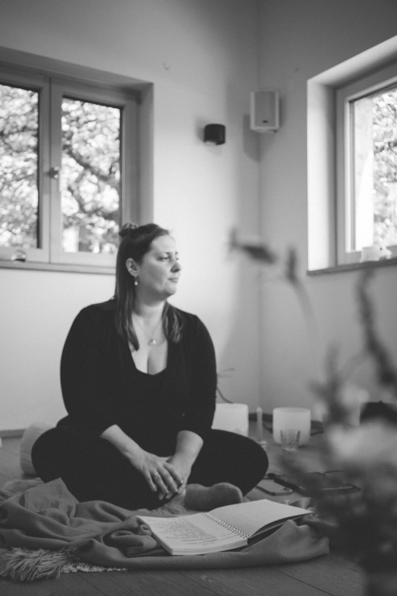 retreat photography - A woman sitting cross-legged on a blanket on the floor, practicing meditation or yoga with her eyes closed and hands resting on her knees, with open books and notes around her in a well-lit room.