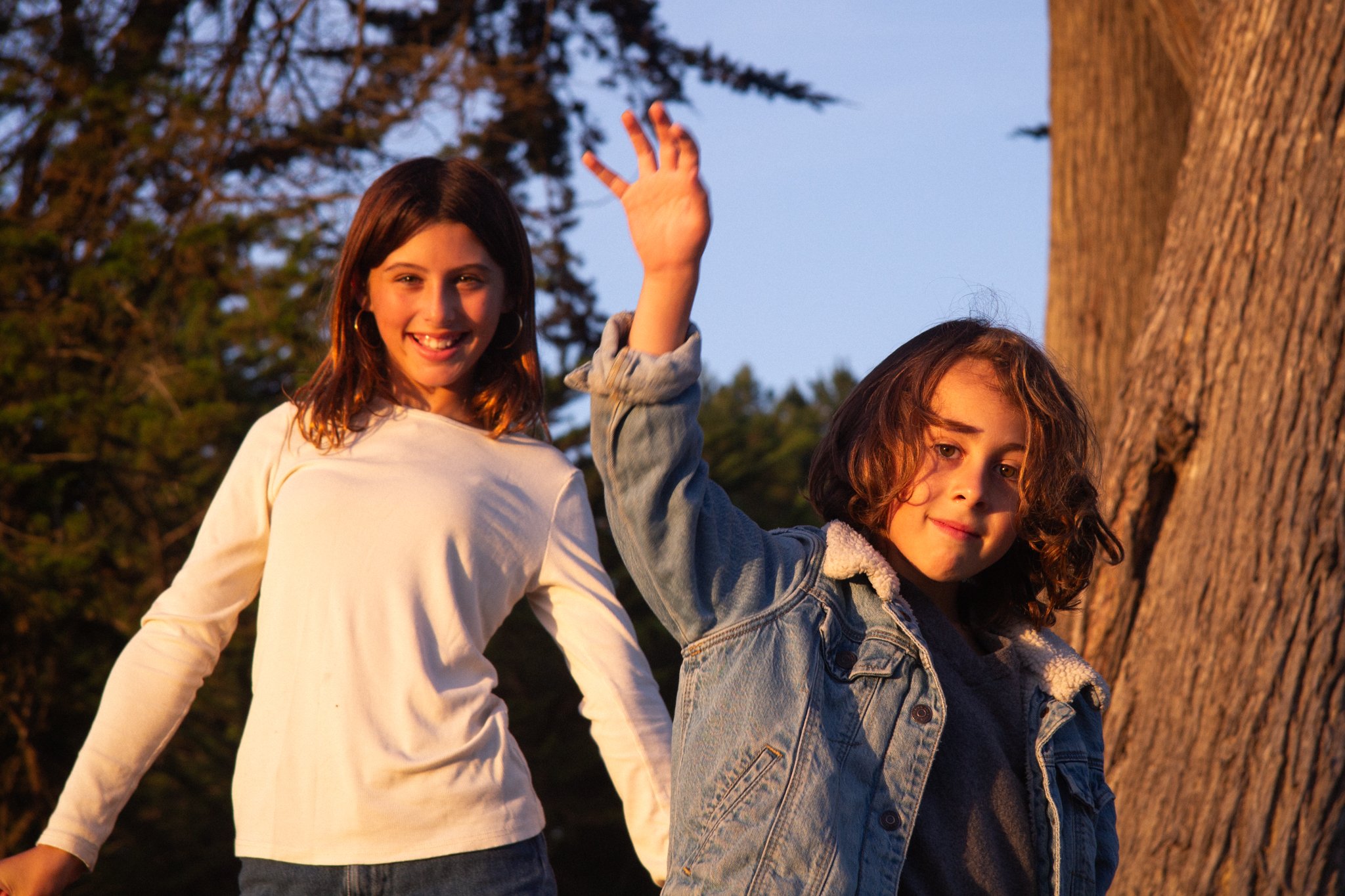 family photo shoot - siblings with brown hair, smiling and waving, standing outdoors near a large tree at sunset.