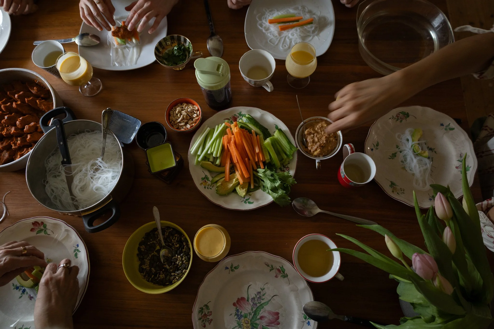 friends photoshoot - A top-down view of a table set with various dishes for a meal, including plates with vegetables, bowls of noodles, sauces, and drinks, with hands reaching for food.
