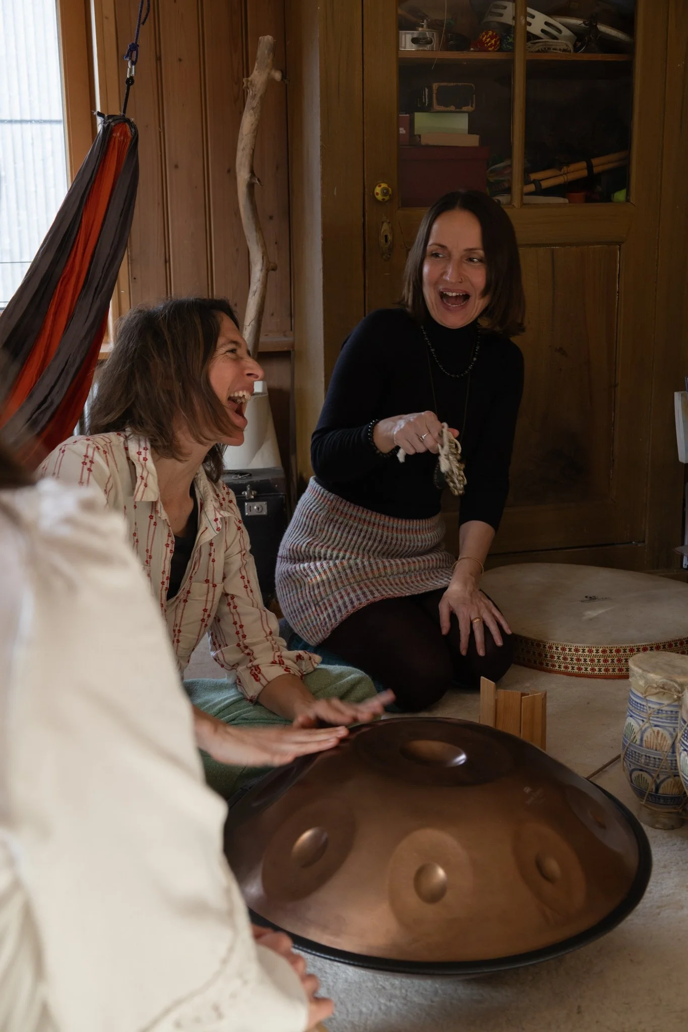 friends photoshoot - Two women share a joyful moment indoors, one sitting and the other standing while holding a musical percussion instrument, with warm wooden walls and shelves in the background.