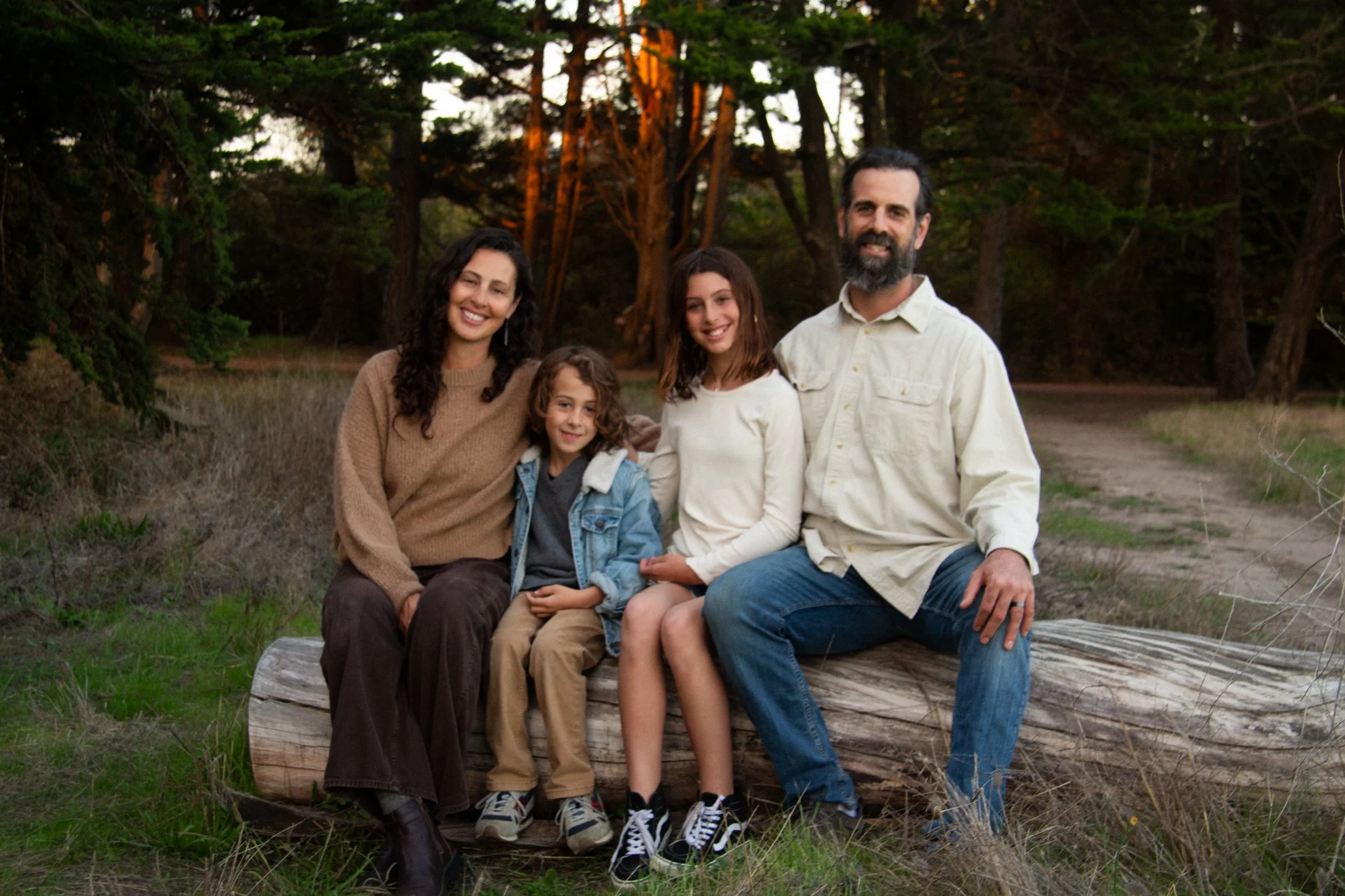 family photoshoot - A family of five sitting on a wooden bench outdoors in a forested area during sunset. The group includes a mother, a father, two kids, smiling and enjoying each other's company.
