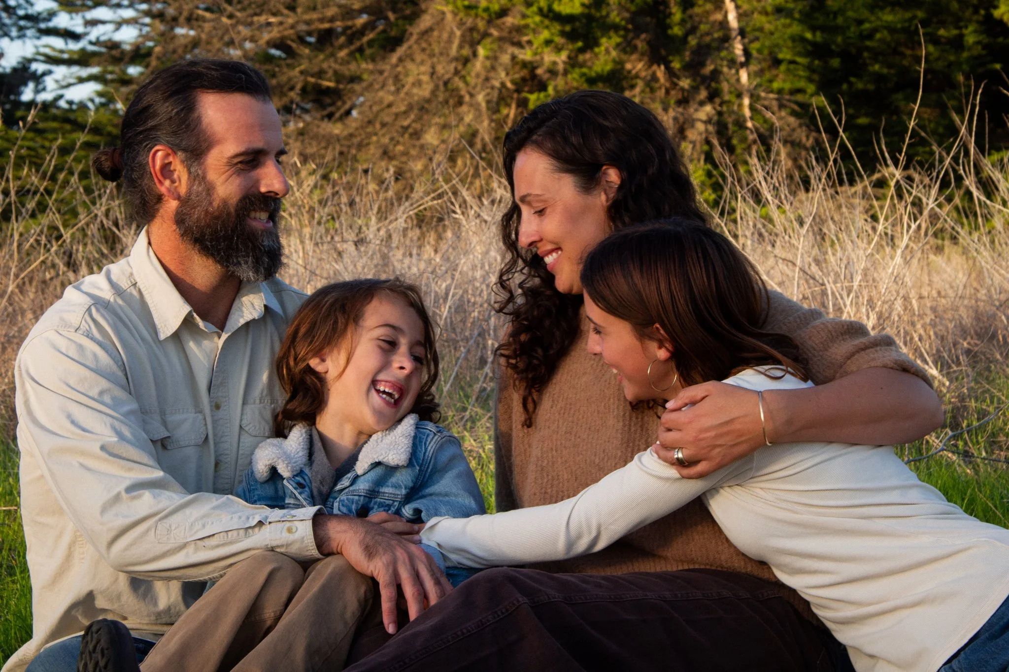 family photo shoot - A family of four sitting outdoors in a grassy area with trees, smiling and sharing a moment of affection.