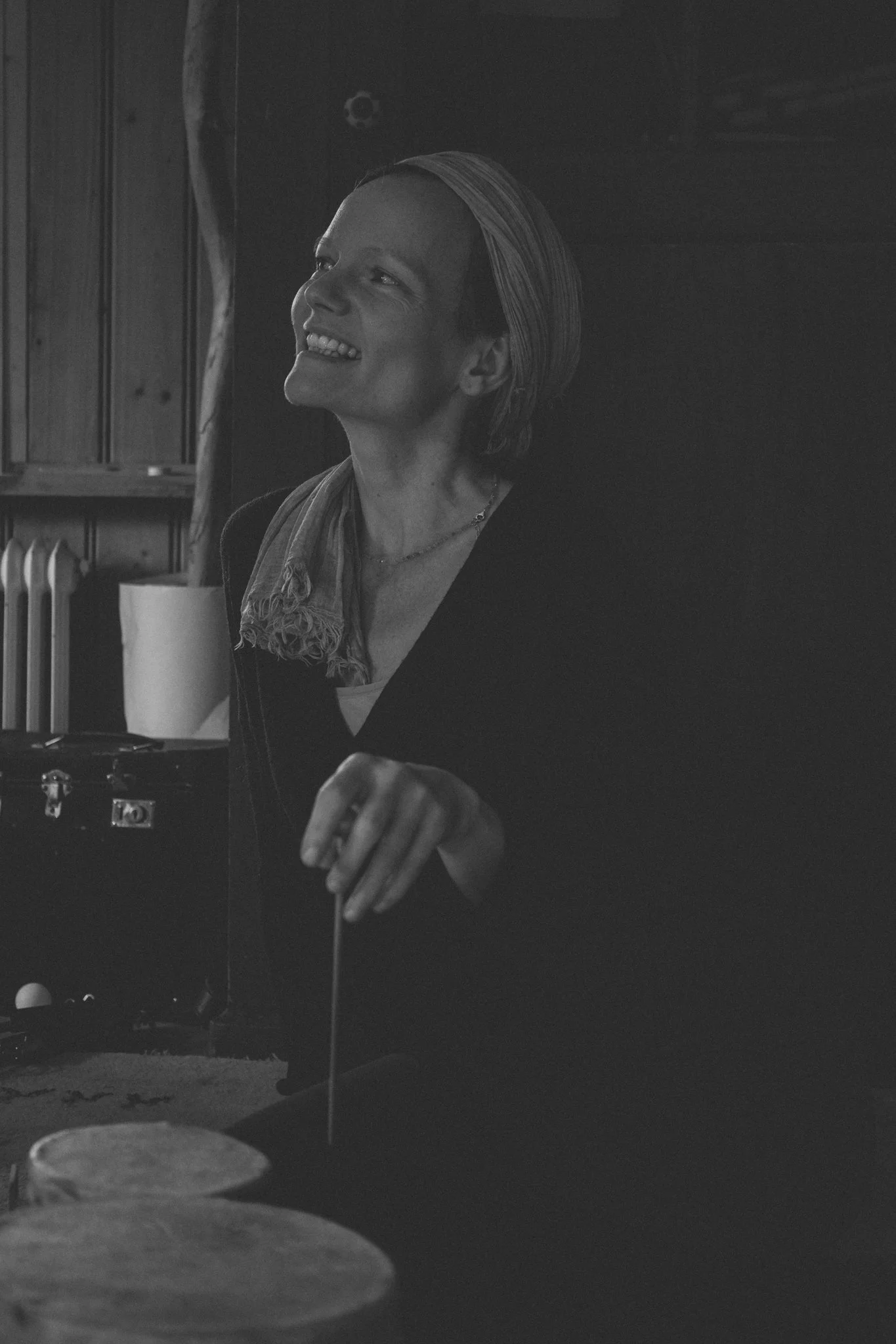 friends photoshoot - A woman with short hair smiling and looking to the side, sitting at a table in a dimly lit room with wood-paneled walls. There are percussion instruments in front of her and a microphone stand nearby.