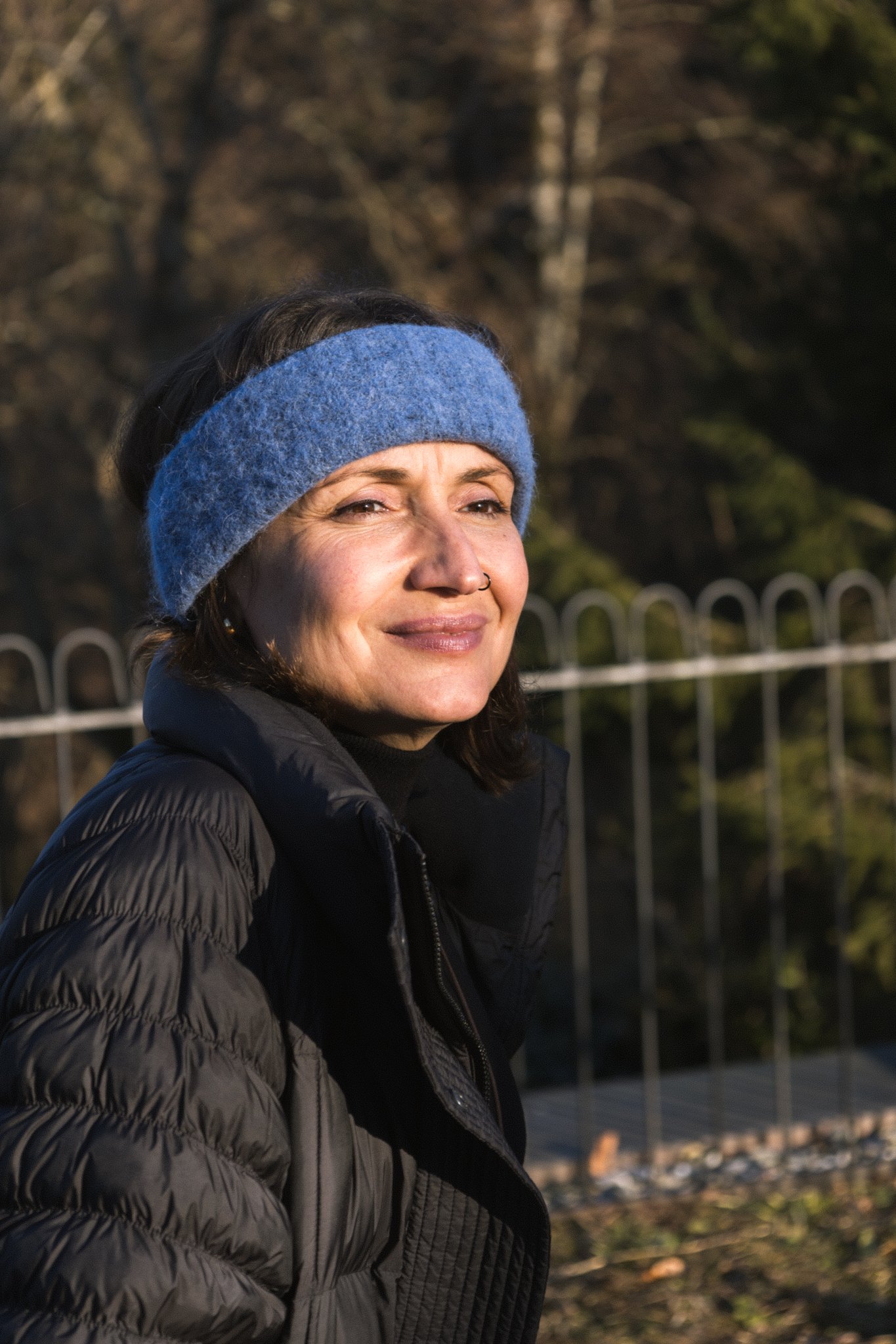 friends photoshoot - A woman wearing a blue headband and a black puffer jacket, outdoors with a metal fence and trees in the background, during daylight.