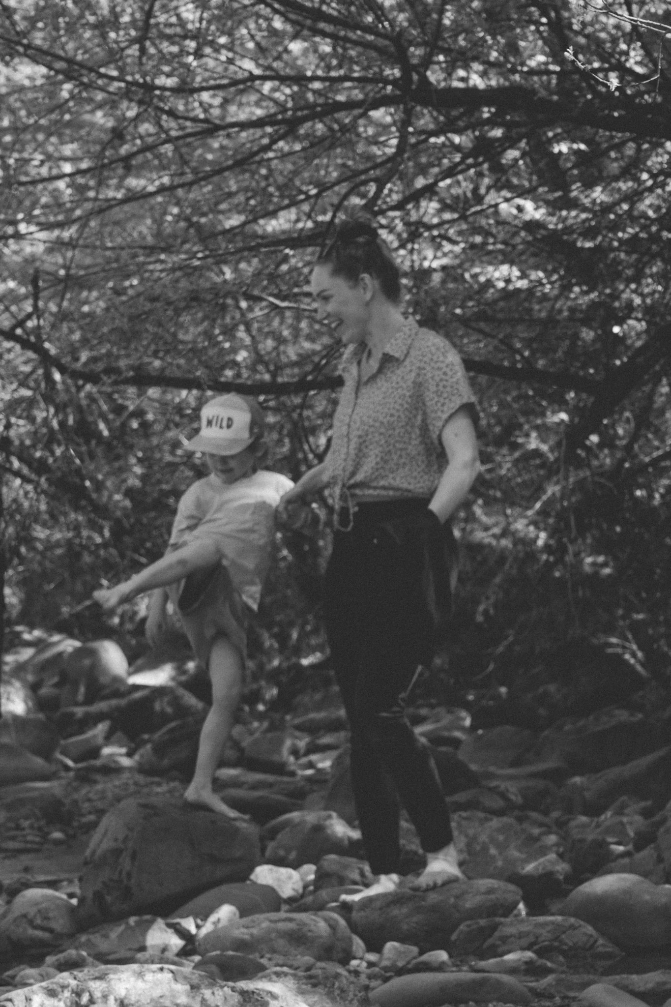 family photoshoot - A mother and a young boy sitting on rocks by a stream in a wooded area, smiling and holding hands.