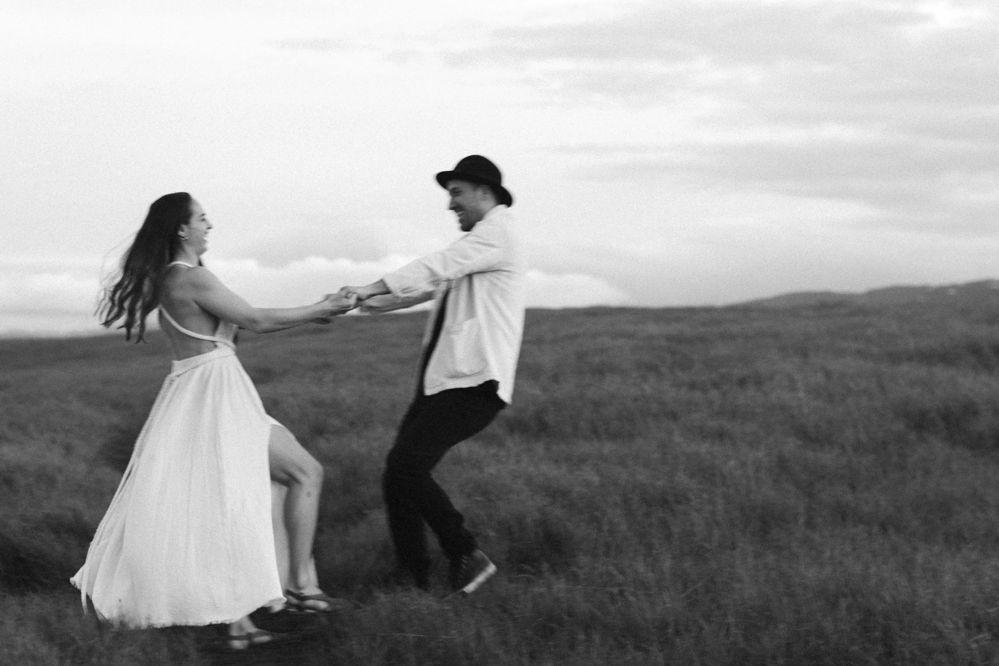 couples photoshoot - A black and white photo of a couple dancing outdoors in a field, holding hands and smiling, with the woman wearing a long white dress and the man wearing a hat.
