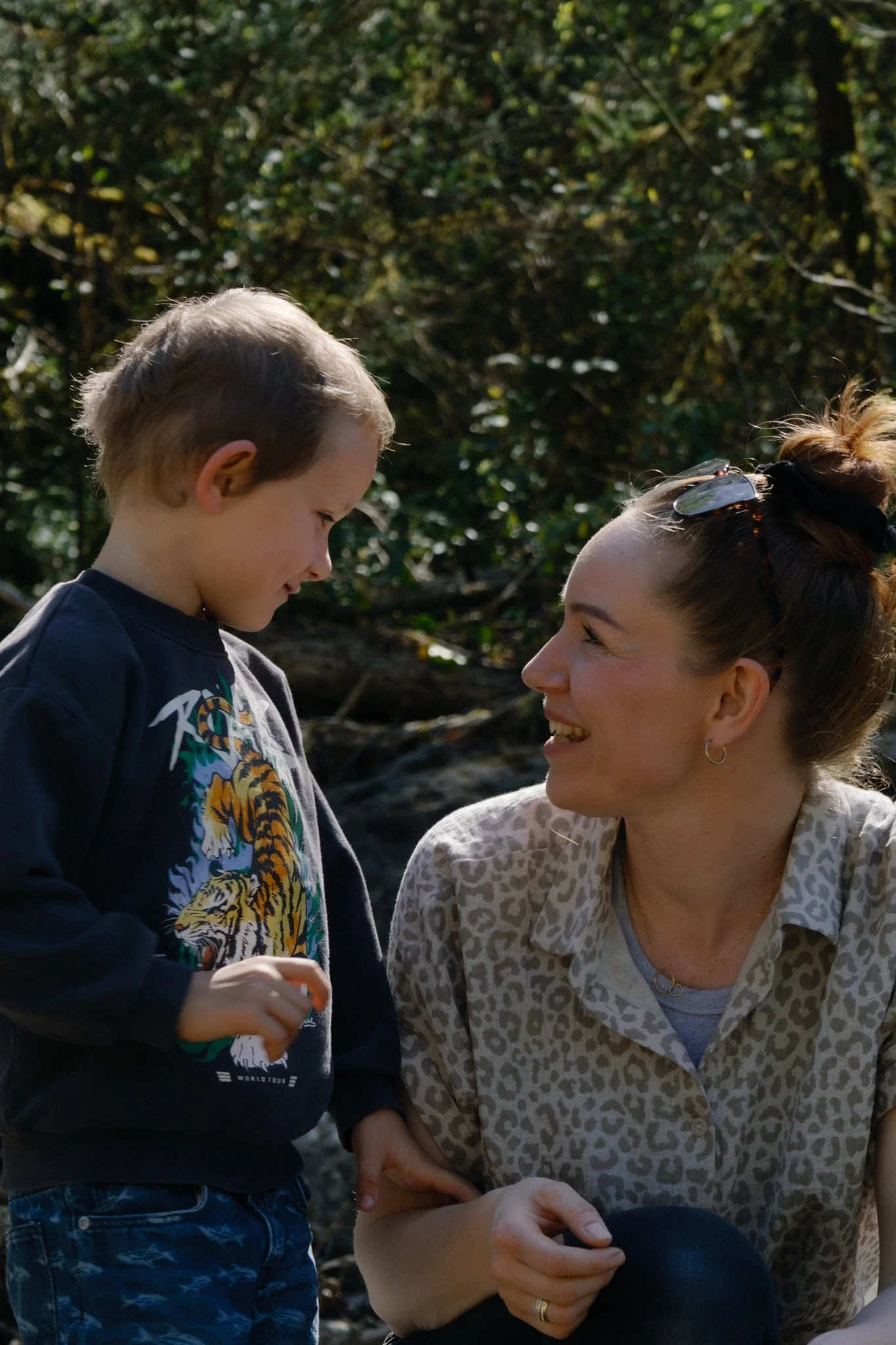 family photoshoot - A mother with sunglasses on her head smiling at a young boy outdoors, both looking at each other with a forest background.