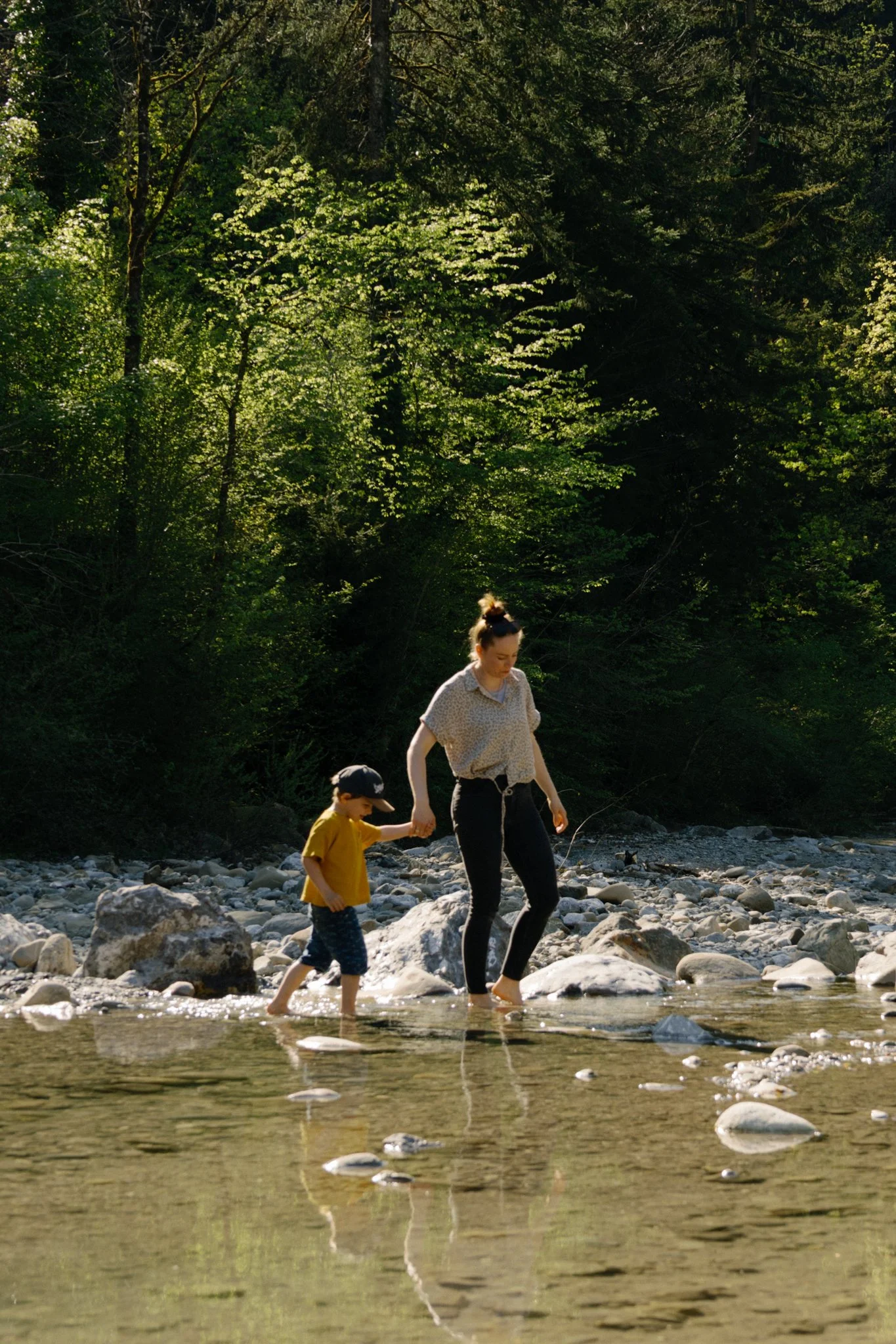 family photoshoot - A mother and her young boy crossing a shallow river with rocky banks and a dense green forest behind them.