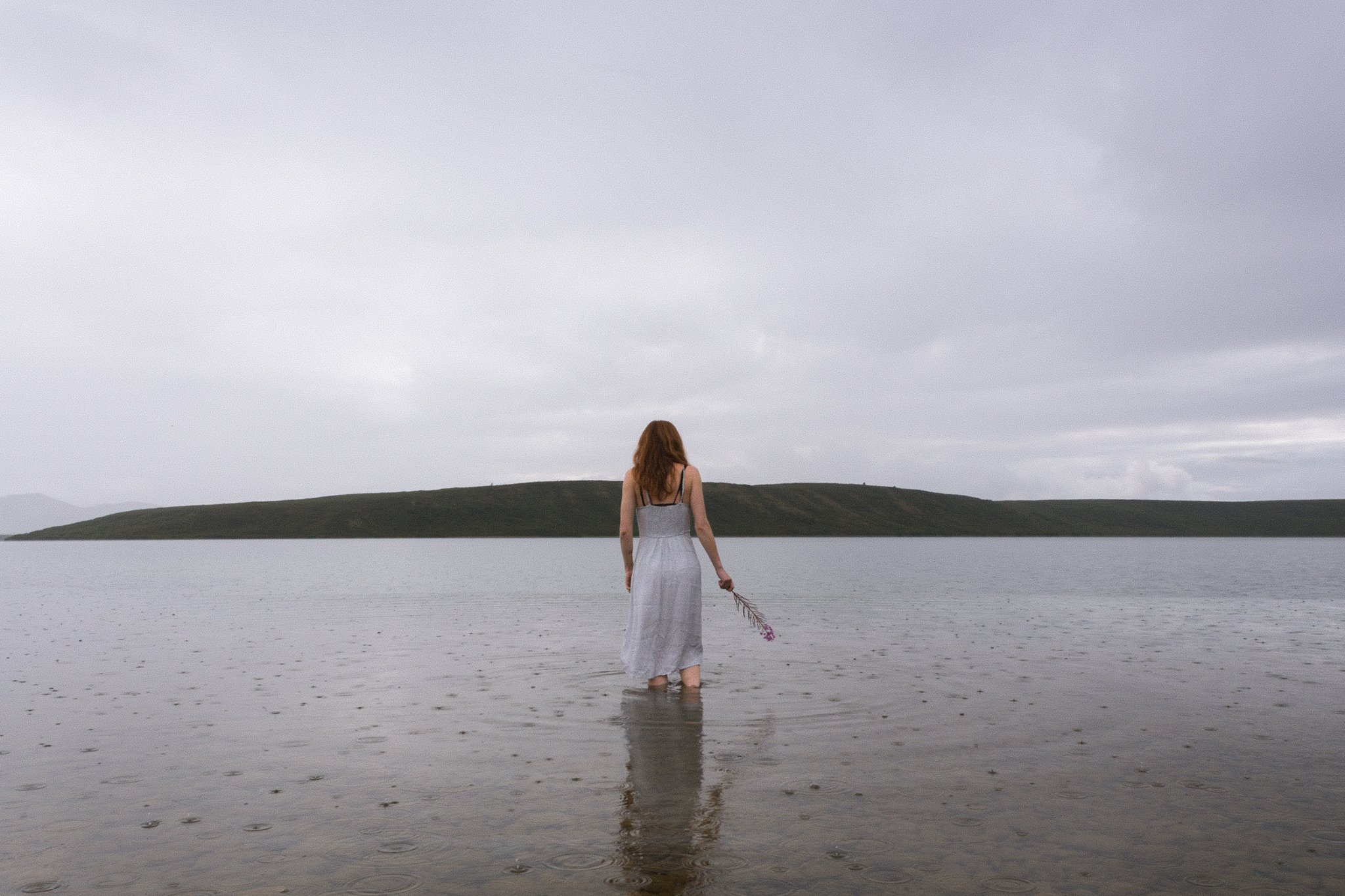 portrait - A woman with long red hair in a white dress stands in shallow water, holding a bouquet of purple flowers, looking towards a large lake with a grassy hill and cloudy sky in the background.