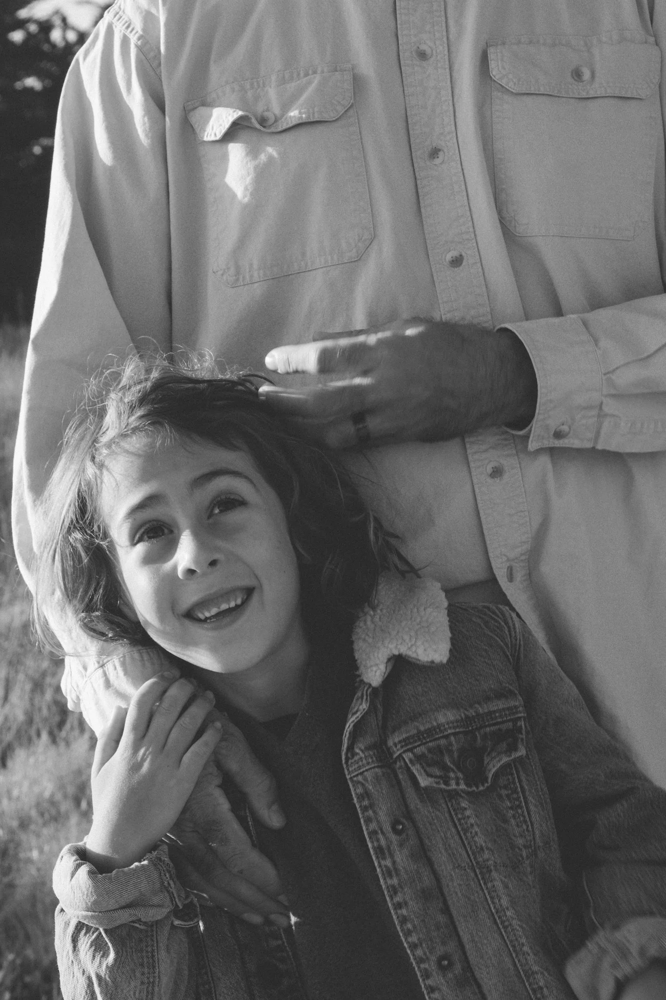 family photo shoot - A young boy with curly hair smiling and looking up, hugging his dad, with both wearing denim jackets and a shirt, outdoor setting with blurred background.