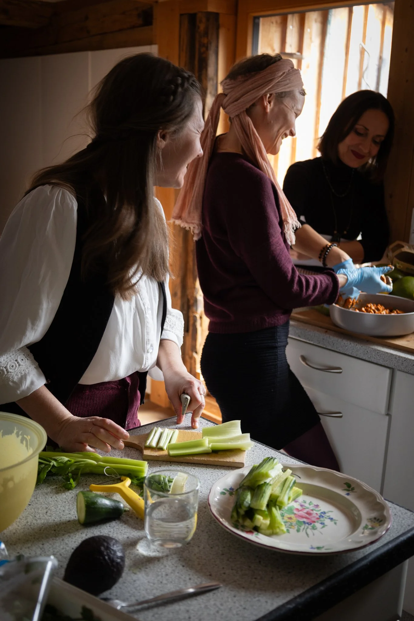 friends photoshoot -Three women preparing vegetables in a rustic kitchen. One woman is slicing celery on a cutting board, while others are involved in food prep, with various vegetables and kitchen tools visible on the countertop.