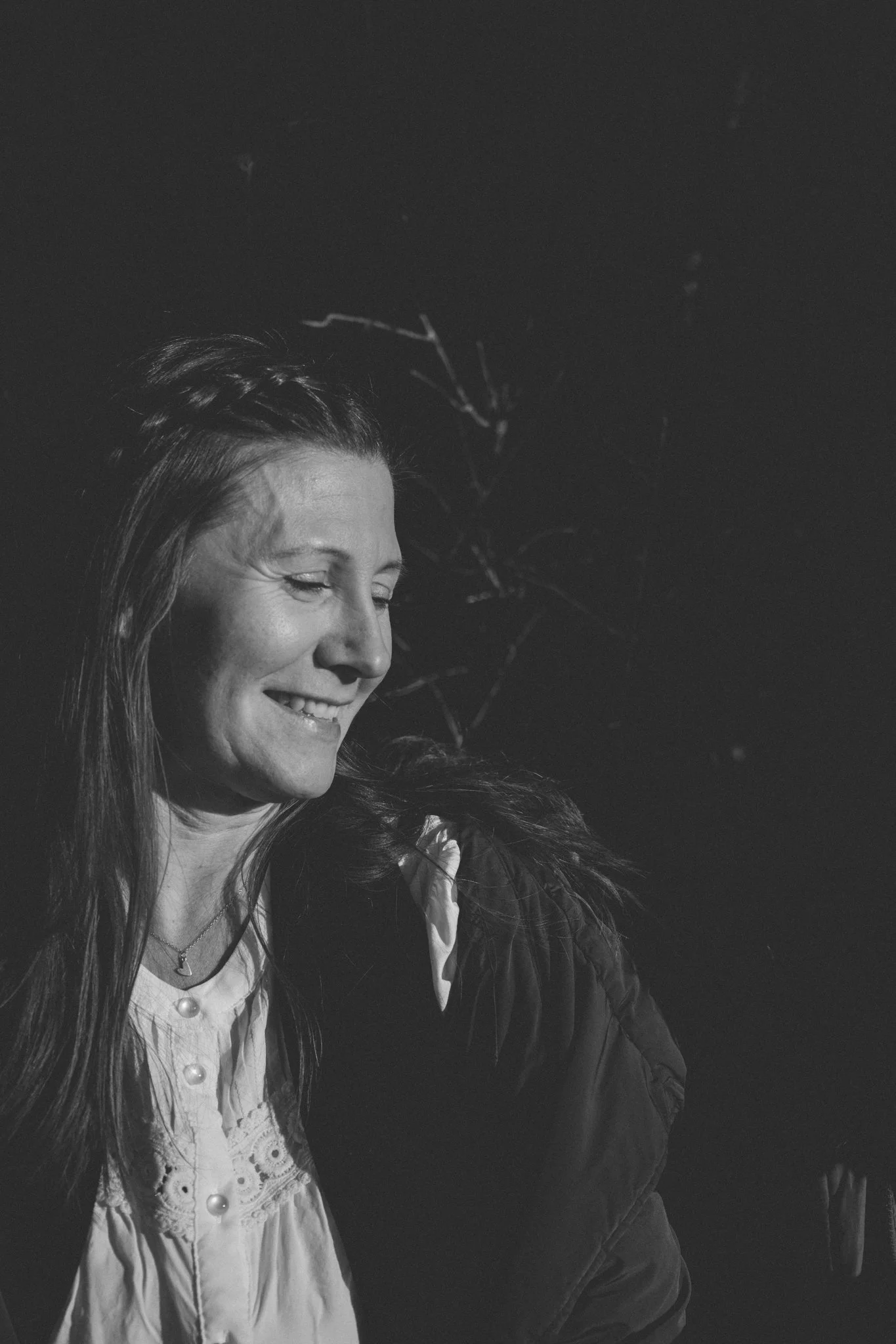 friends photoshoot - A smiling woman with long hair in a white blouse and a necklace, standing outdoors with a dark sky and leafless branches in the background.