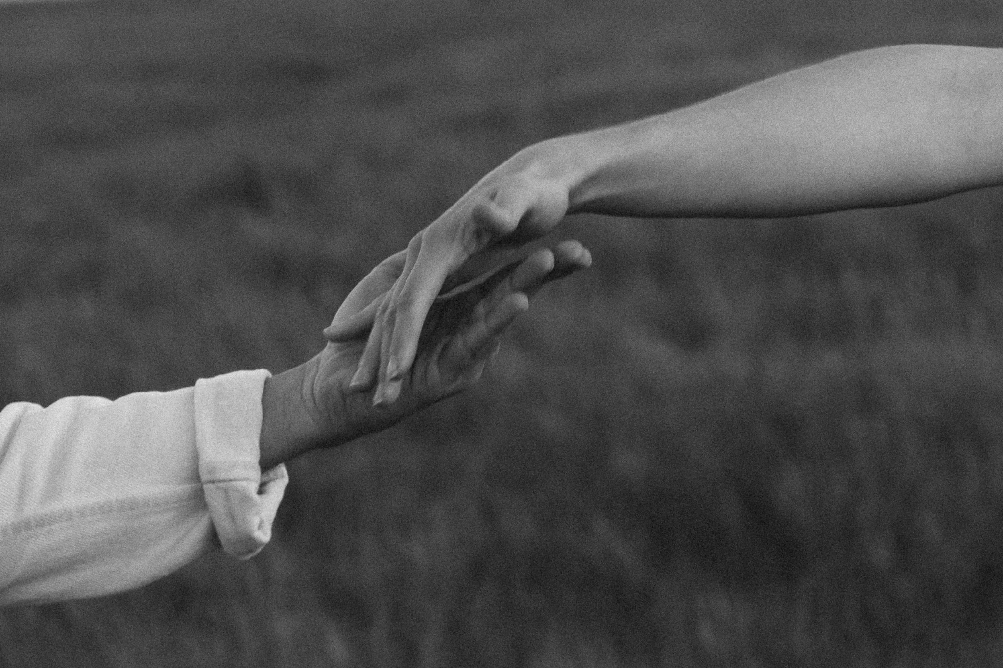 couples photoshoot - Close-up of two hands gently touching against a dark background, one hand appears to be resting over the other, in black and white.