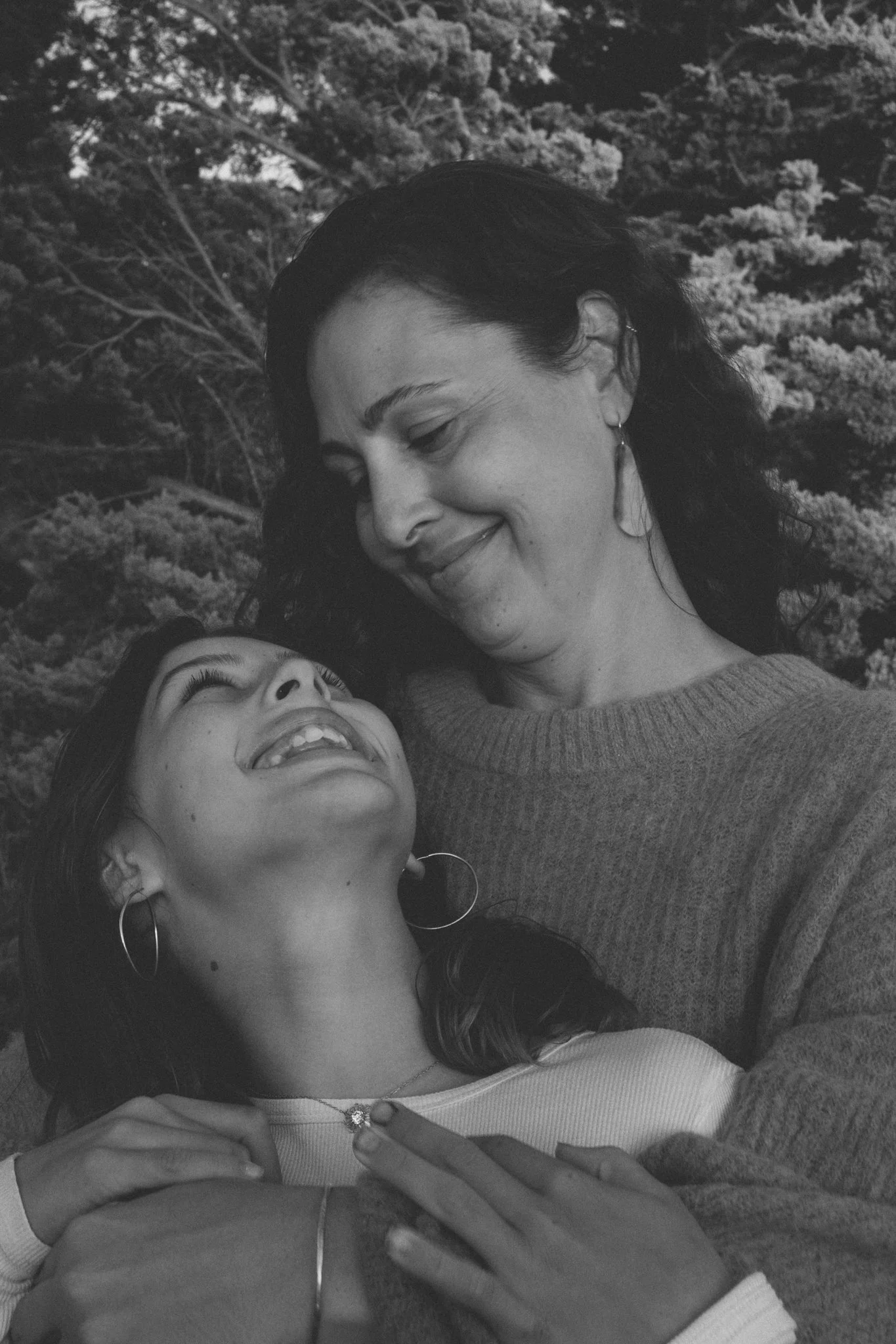 family photo shoot - a mother and her daughter, smiling and looking at each other. The background is an outdoor setting with trees.