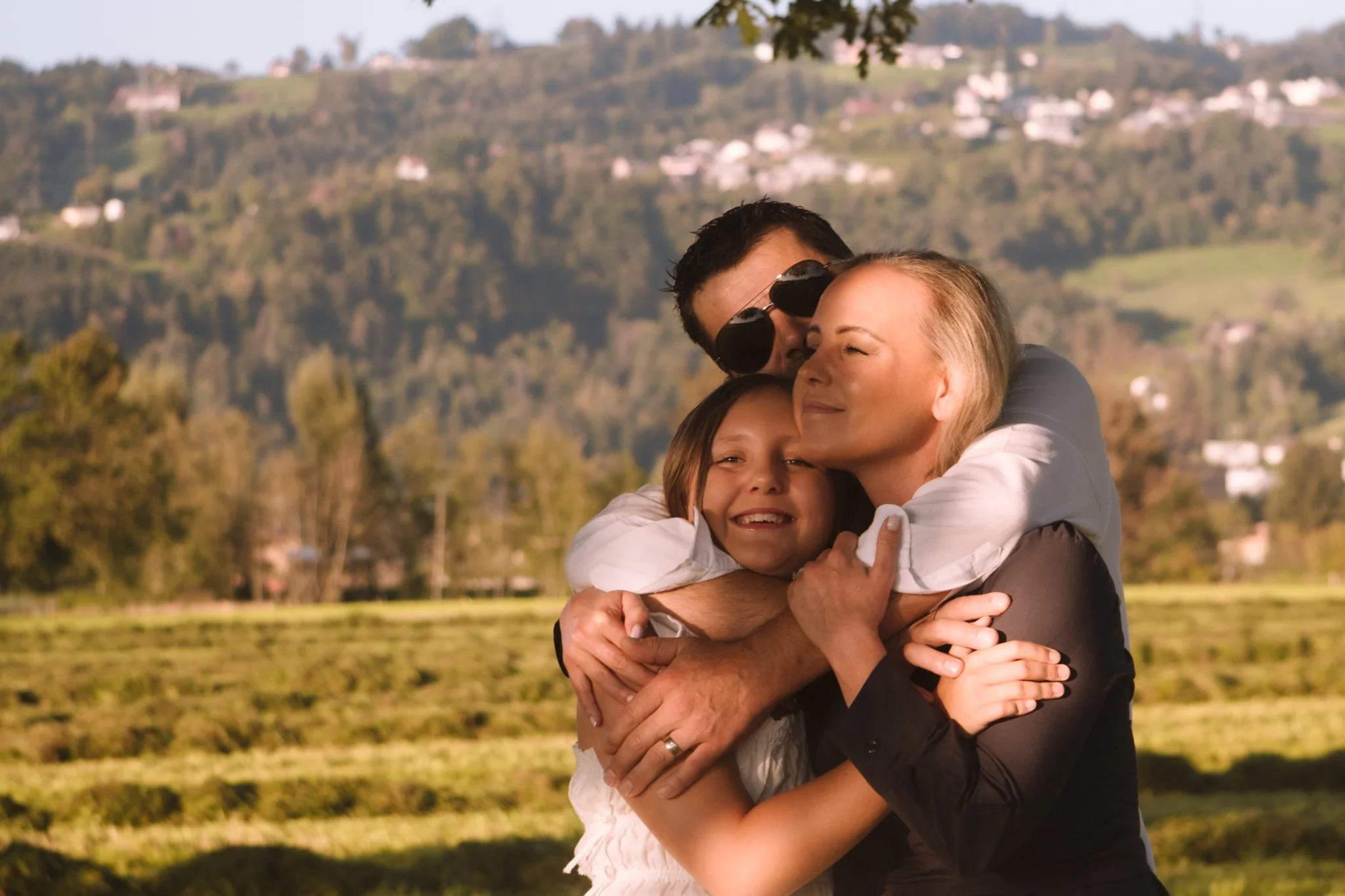 family photoshoot - A family hugging outdoors with green fields, trees, and houses on hills in the background.