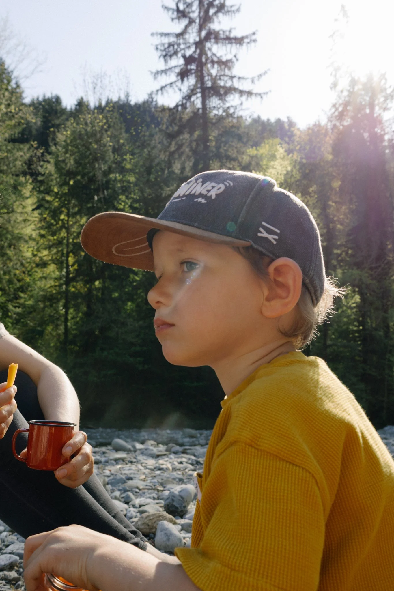 family photoshoot - A young boy with blonde hair wearing a gray cap with white text and a yellow shirt, sitting outdoors near a rocky riverbank with trees and sunshine in the background.