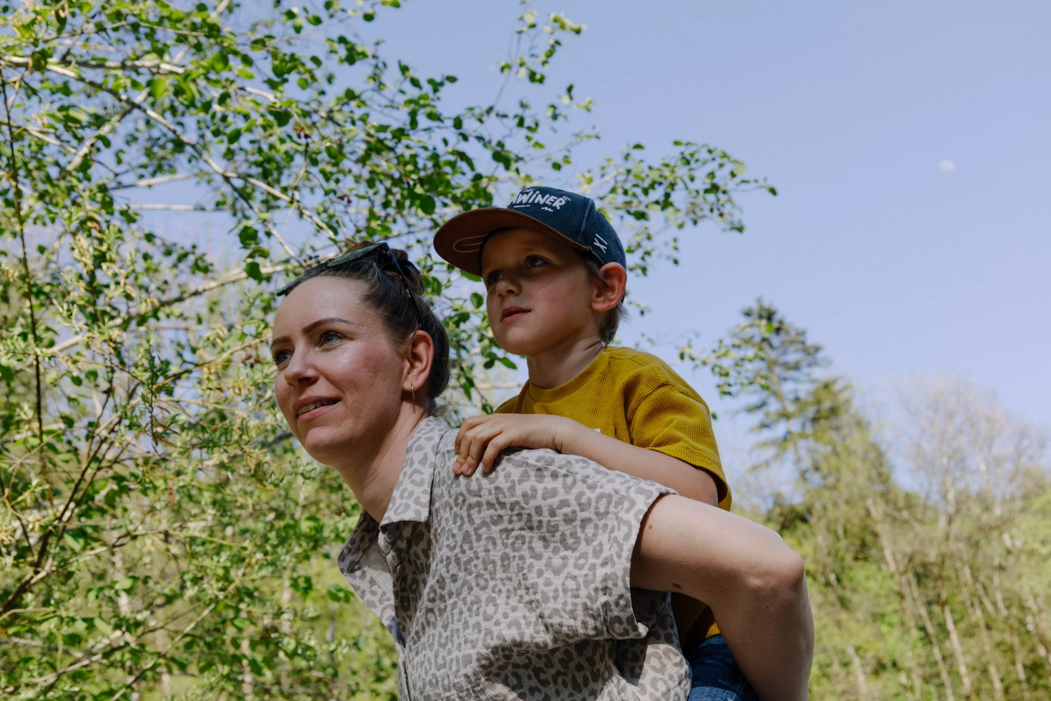 family photoshoot - A woman with brown hair and a patterned gray shirt is giving a piggyback ride to a young boy with a baseball cap and a yellow shirt outdoors, with green trees and a blue sky in the background.