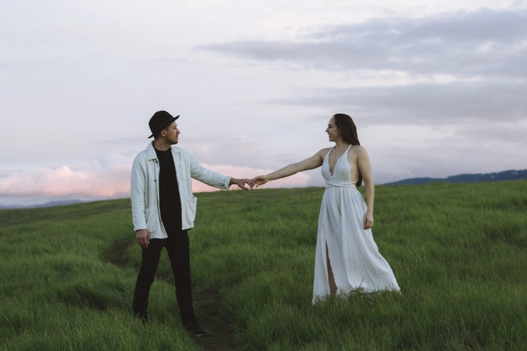 couples photoshoot - A man wearing a black hat and light jacket holding hands with a woman in a white dress standing on a grassy field under a cloudy sky.