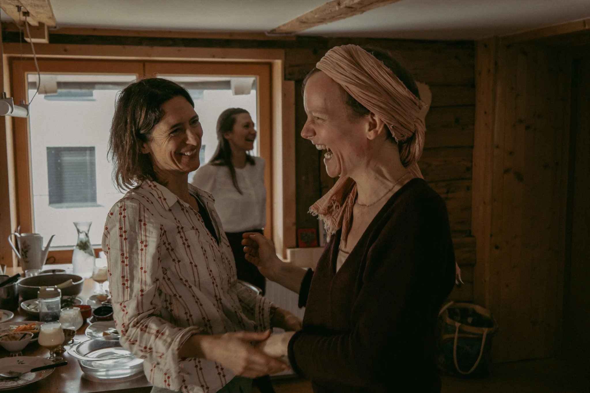 friends photoshoot - Two women smiling and holding hands, one with dark hair and the other with a headscarf, in a cozy wooden kitchen with another woman in the background.