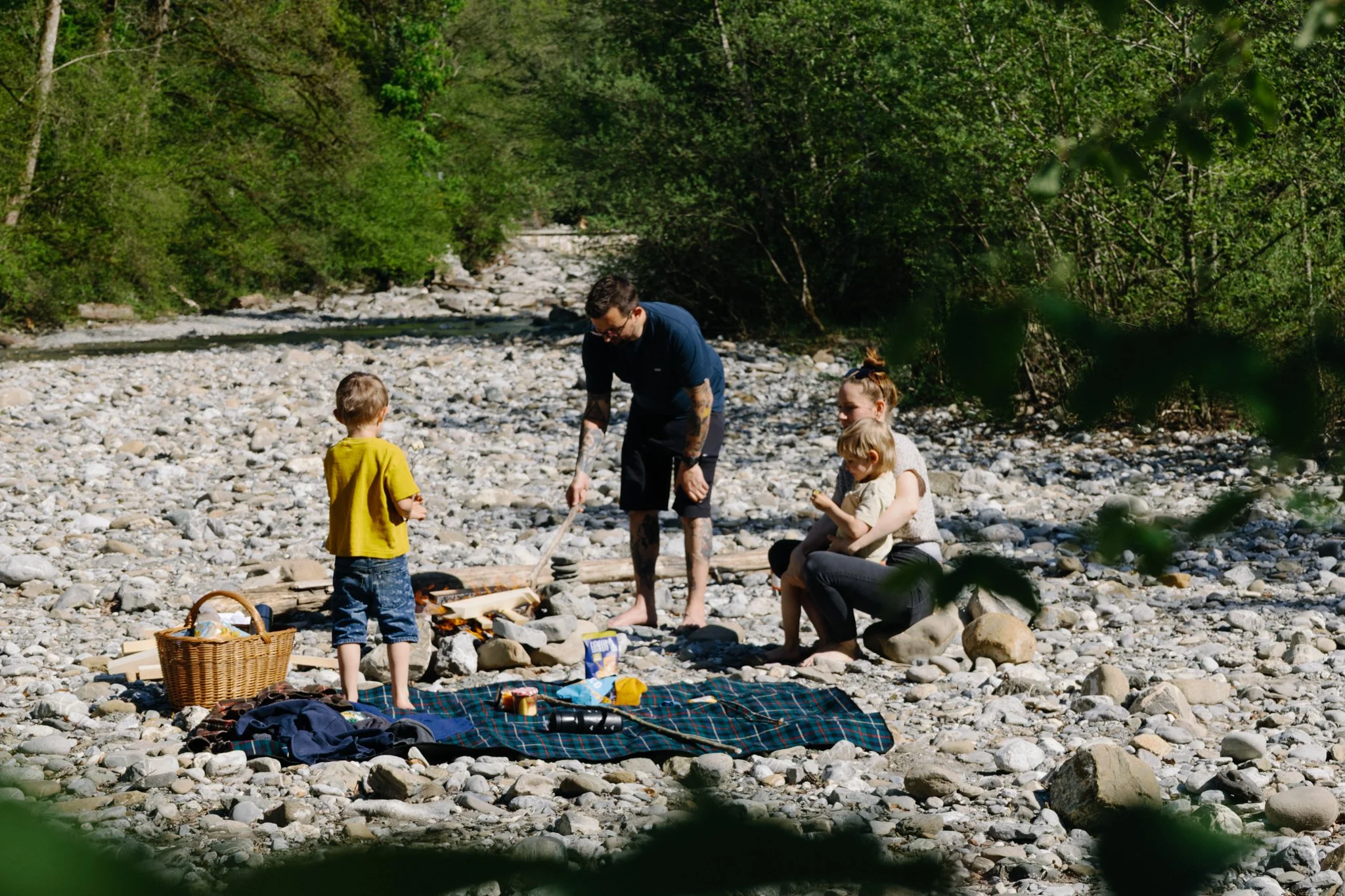 family photoshoot - A family of four enjoying a picnic by a rocky river, surrounded by lush green trees, with one man and one woman and two boys standing.