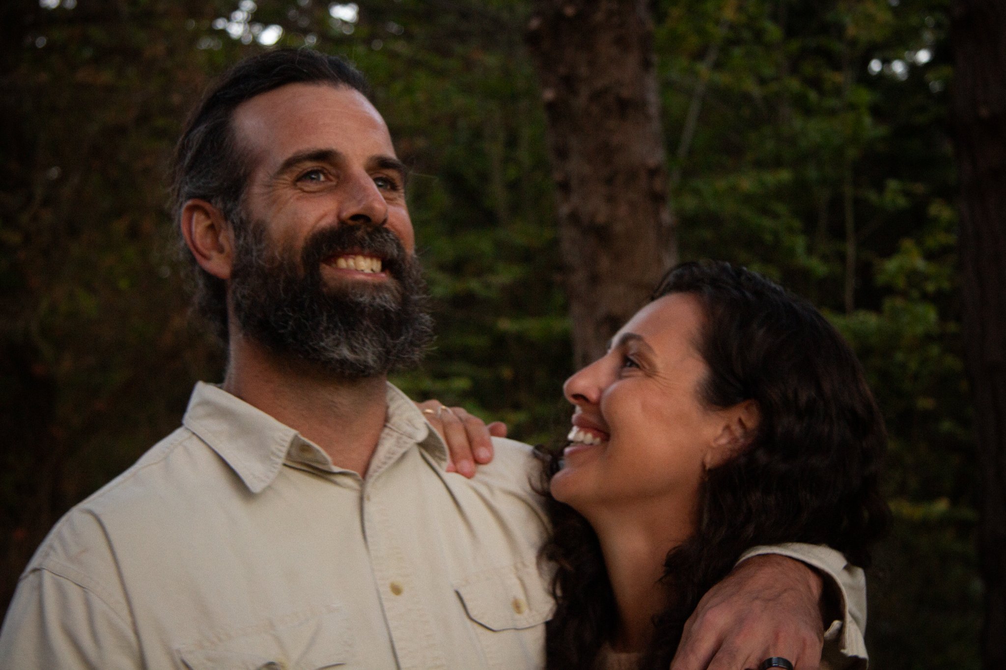 family photo shoot - A smiling man with a beard and a woman smiling, embracing each other outdoors with trees in the background.