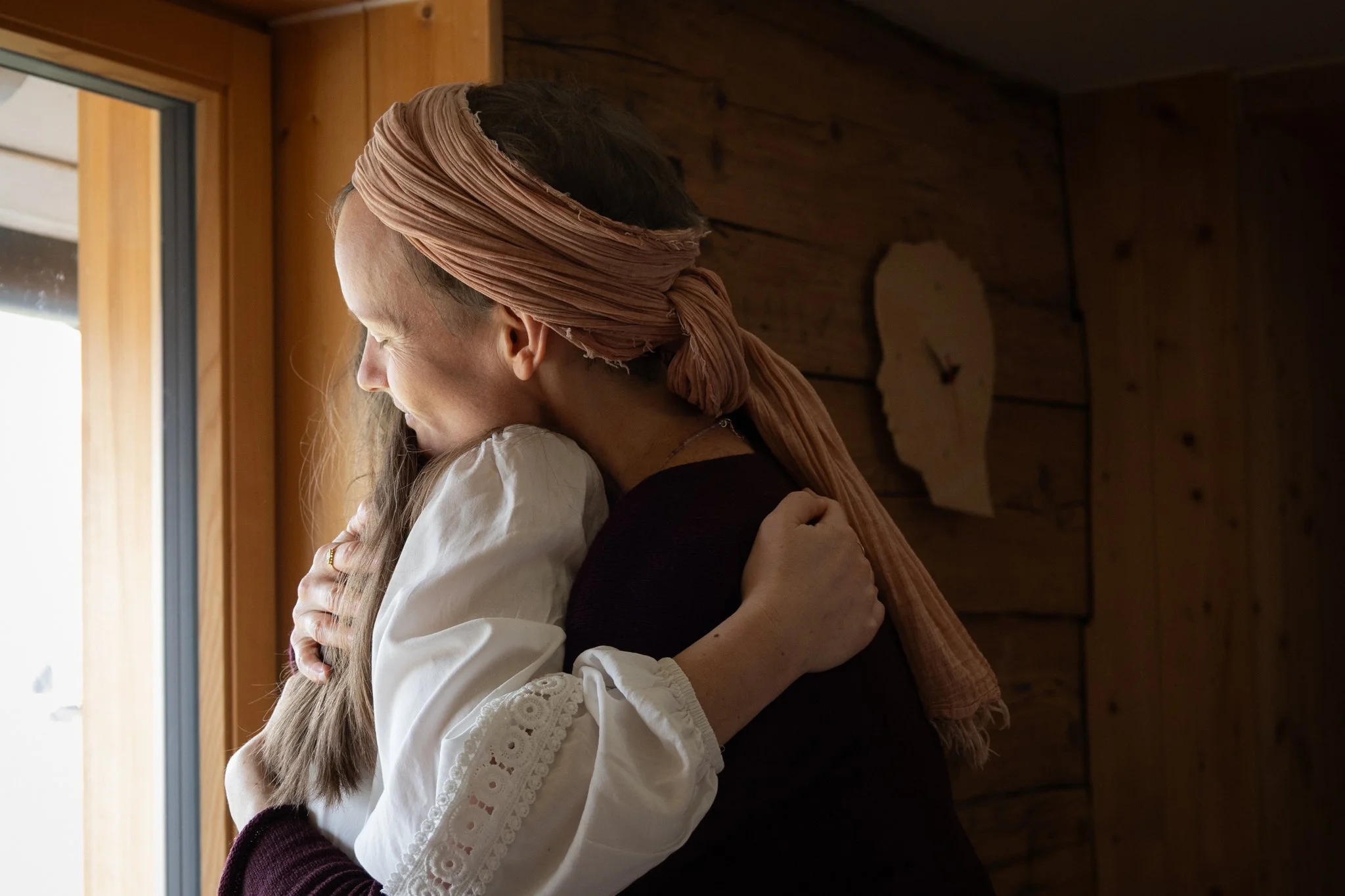 friends photoshoot - A woman with a peach-colored headscarf embraces a girl with long brown hair, near a wooden wall and window in a cozy indoor setting.