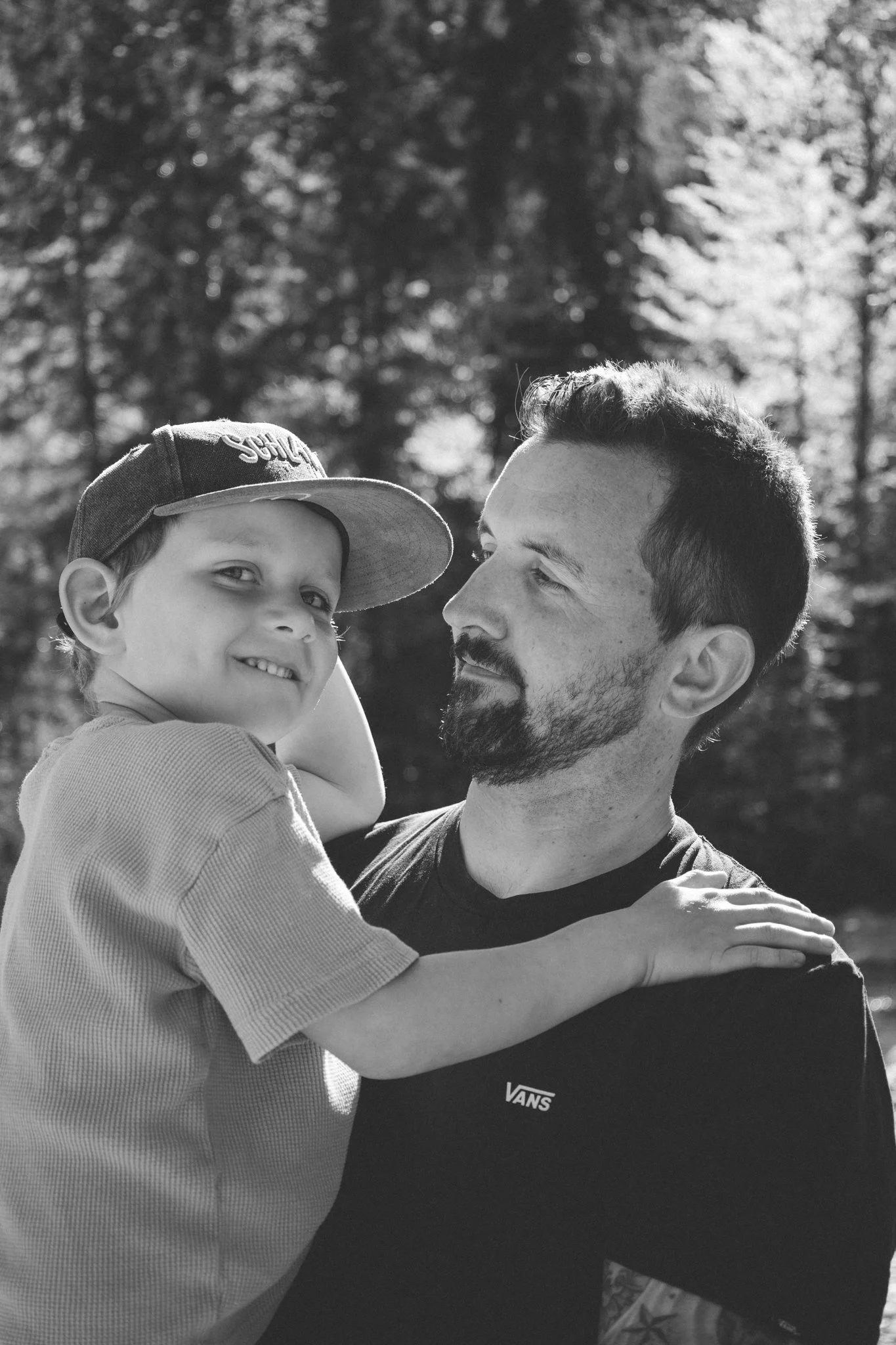 family photoshoot - A young boy wearing a baseball cap and a smiling expression is being held by an adult male. The adult is looking at the boy with a gentle expression. The scene is outdoors with trees in the background, black and white