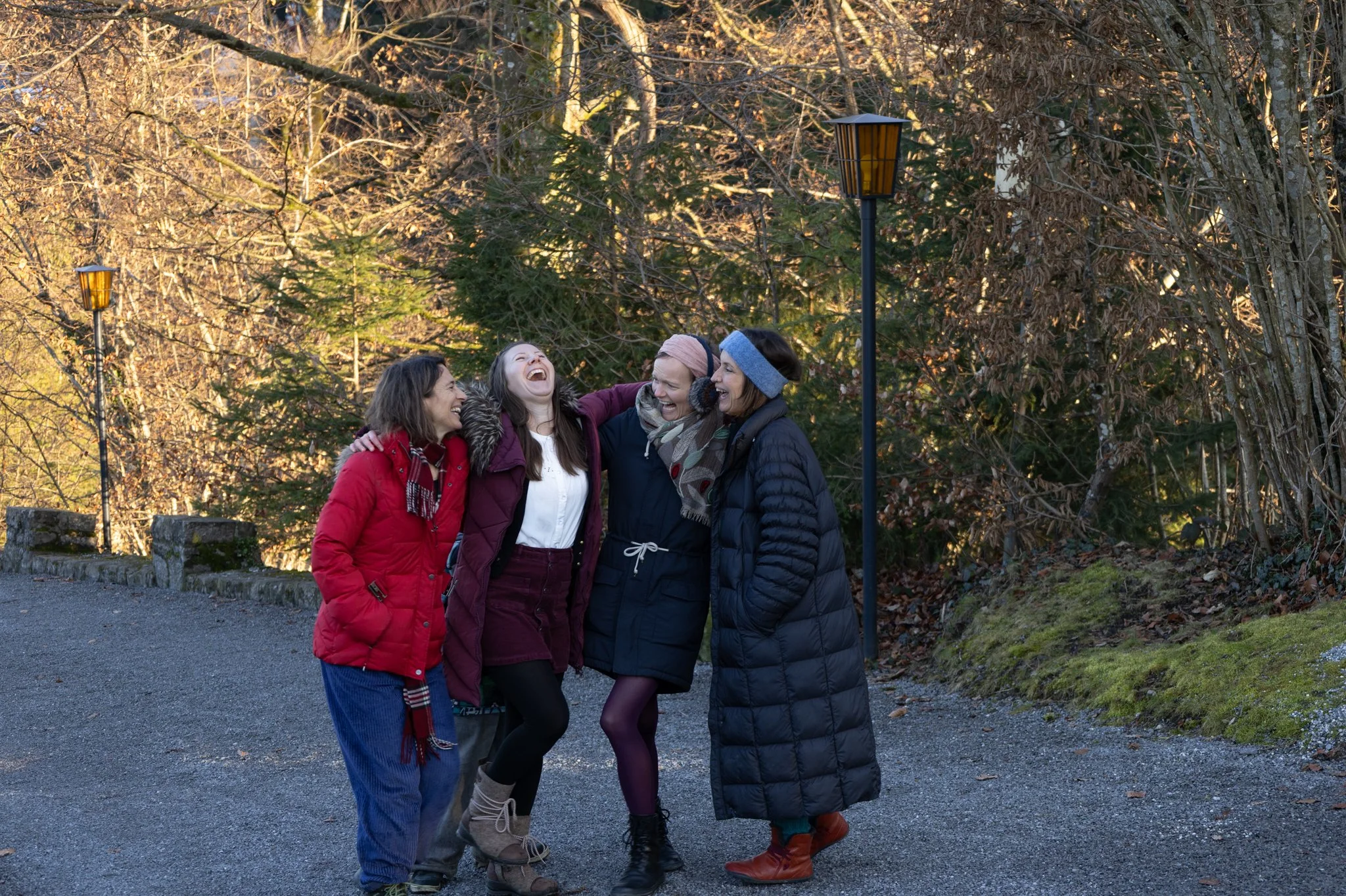friends photoshoot - Four women in winter clothing standing close together outdoors, laughing and smiling with their arms around each other, with trees and street lamps in the background.