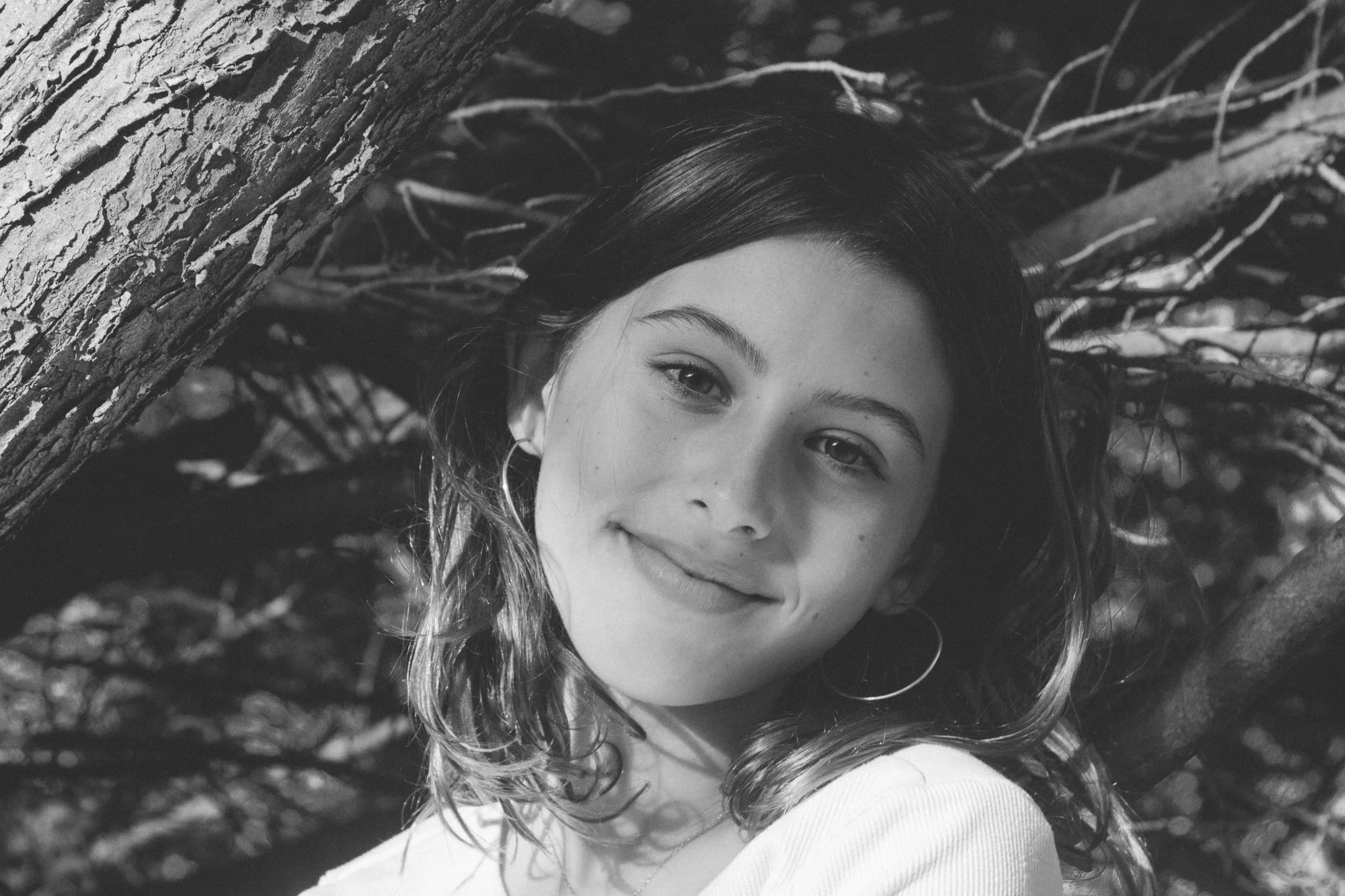family photo shoot - Black and white photo of a young girl with wavy hair, smiling, with a tree trunk and branches in the background.