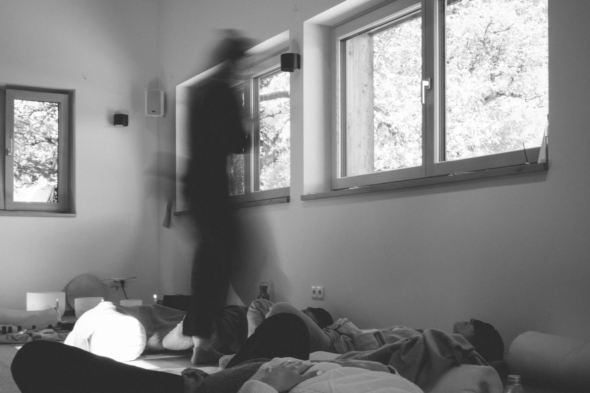 retreat photography - People lying on beds in a room with large windows showing trees outside, and a person standing near the windows, blurred, in a black shirt.
