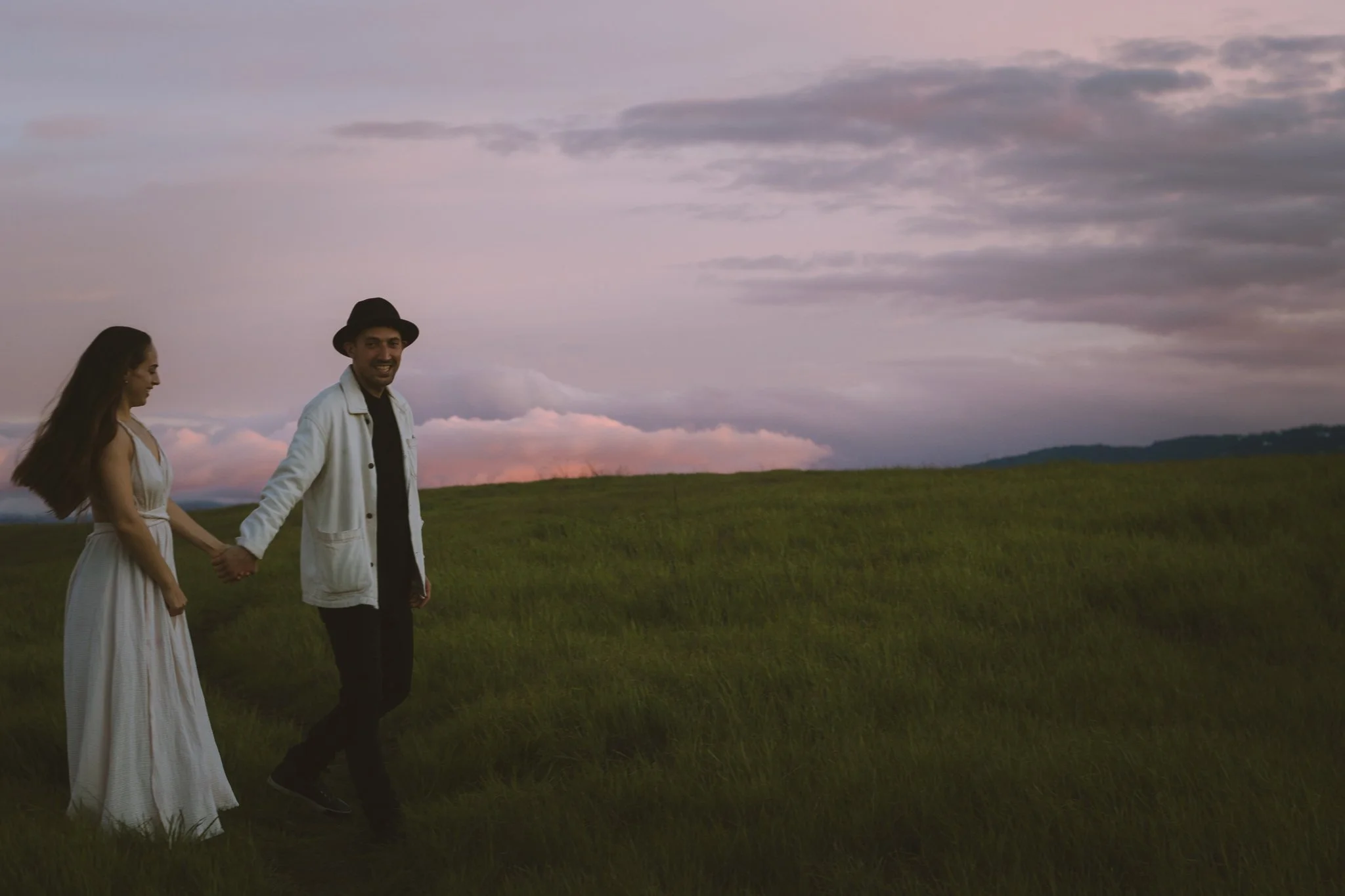 couples photoshoot - A couple holding hands and walking in a grassy field during sunset, with a cloudy sky in the background.