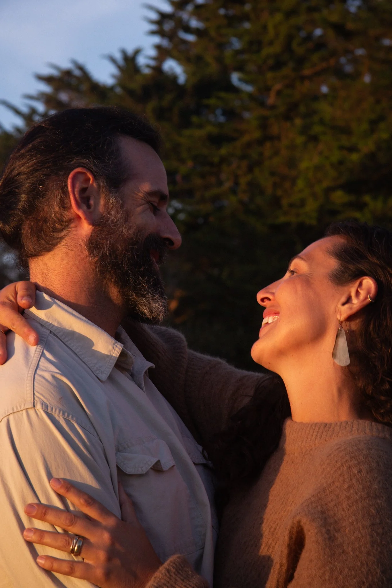 family photo shoot - A man and woman smiling and looking at each other outdoors during sunset, with trees in the background.