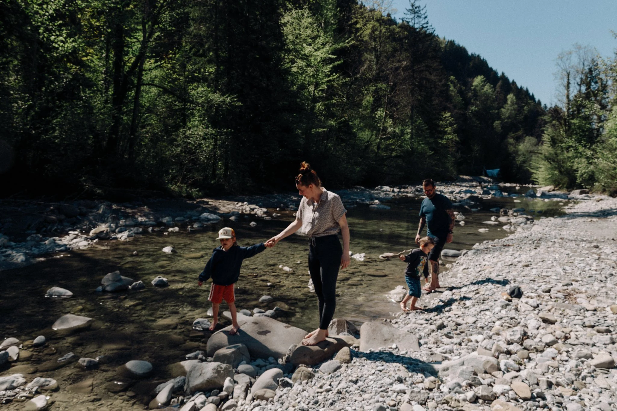 family photoshoot - Family of four crossing a shallow rocky river in a forested area on a sunny day.
